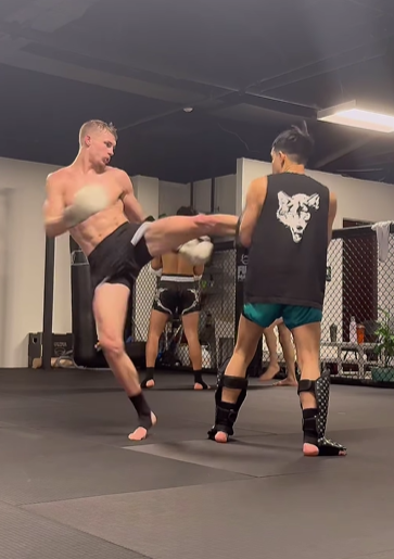 Two men practicing martial arts in a gym with one delivering a high kick to the other's midsection while the other blocks it.