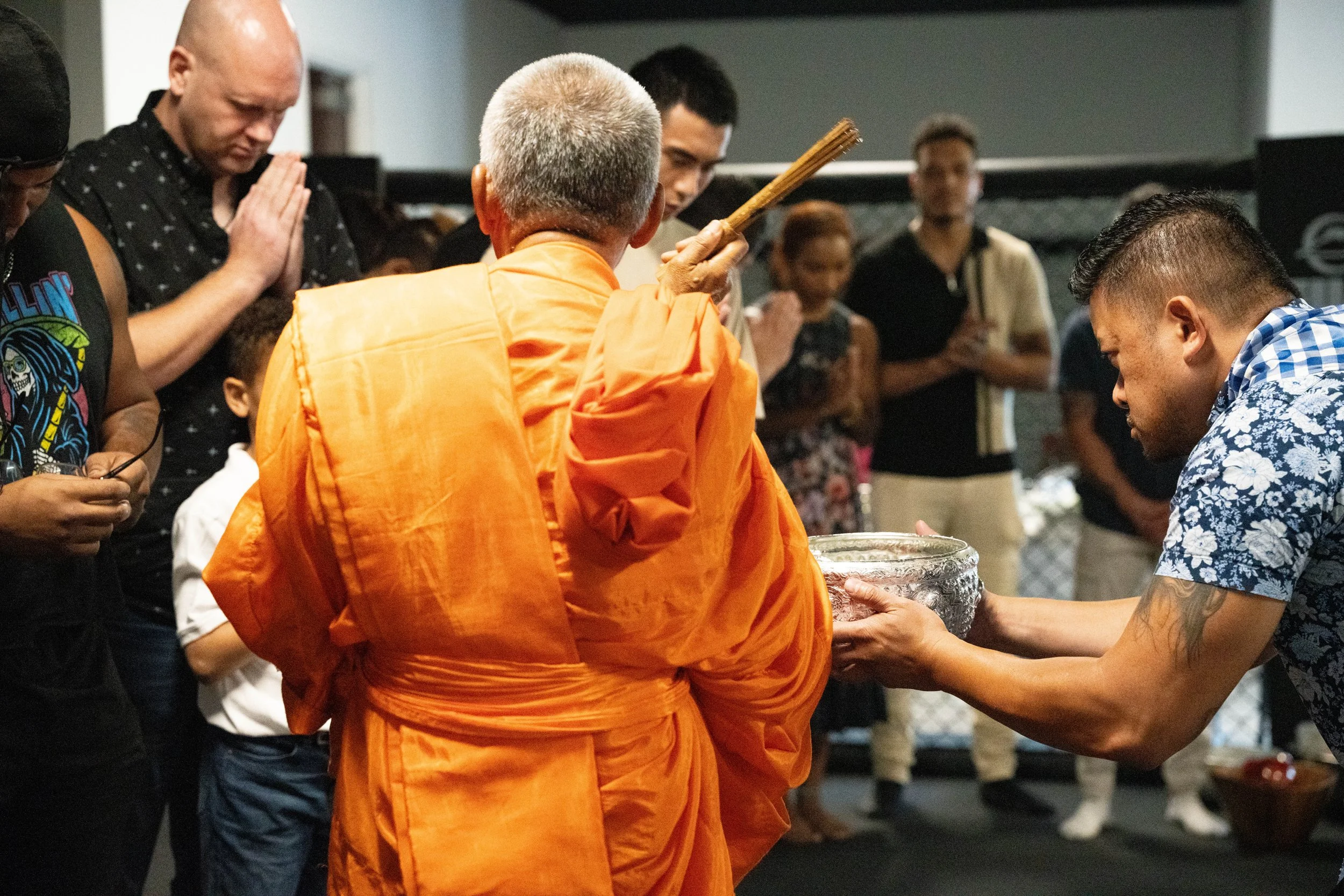A Buddhist monk dressed in an orange robe is holding a stick while receiving a silver bowl from a young man with tattoos on his arm and a blue floral shirt. Several people are standing in the background with prayerful hands and attentive expressions, participating in a religious or spiritual ceremony inside a room.