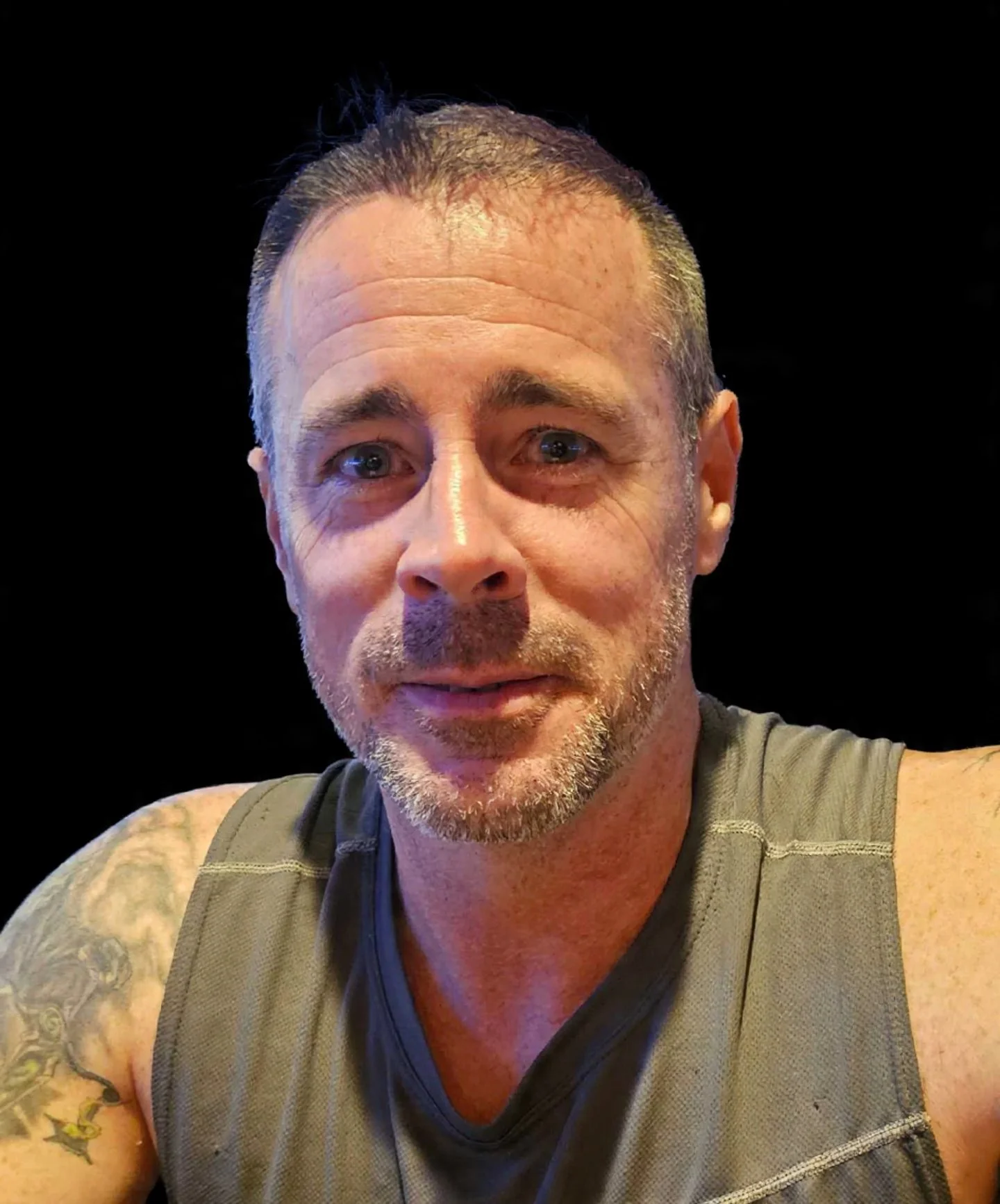 Close-up photo of a middle-aged man with short hair, a beard, wearing a sleeveless shirt, with a tattoo visible on his shoulder, against a black background.