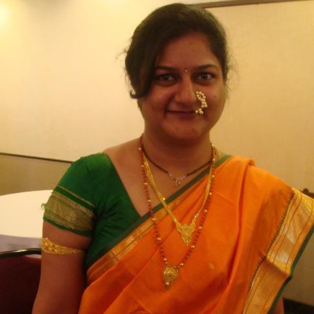 Woman with traditional Indian attire, wearing a green blouse and orange sari, adorned with gold jewelry and a nose ring, smiling indoors.