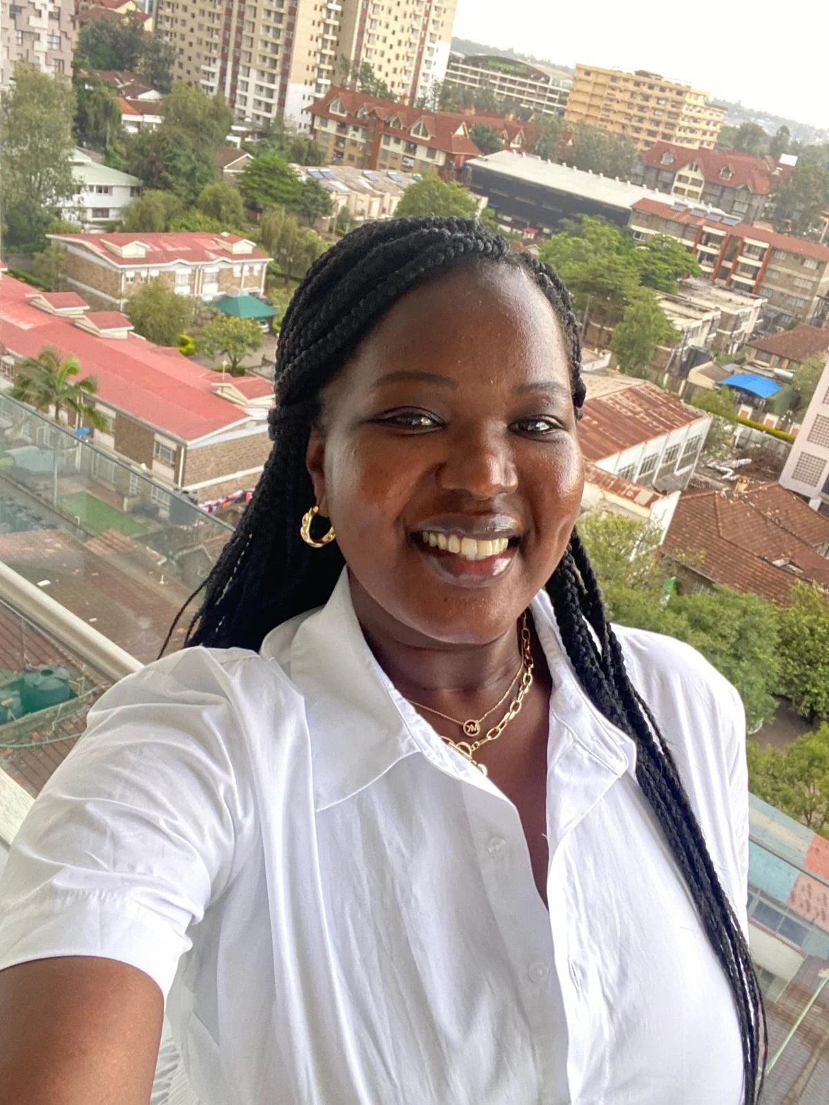 A woman smiling on a balcony with a cityscape background of high-rise and low-rise buildings and trees.
