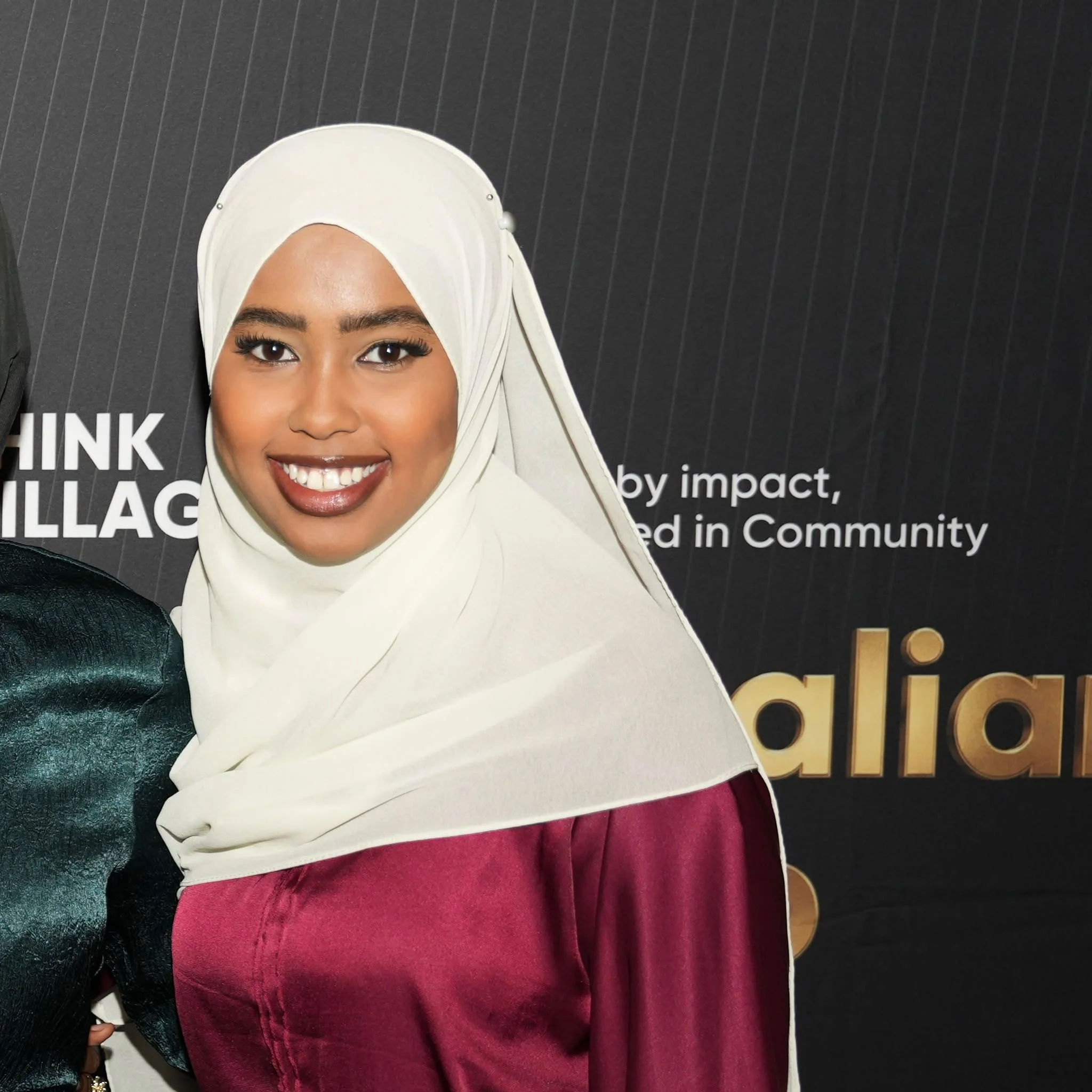 Close-up of a woman wearing a white hijab, smiling, with black hair, dark skin, and red velvet pants, standing against a dark background with white and gold text.