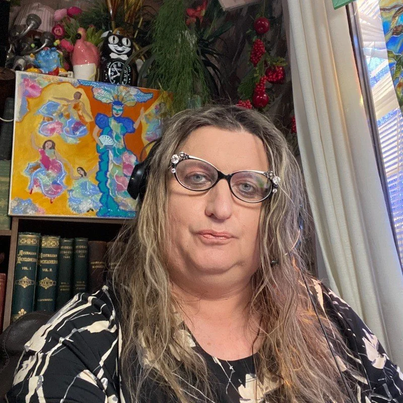 A woman with wavy blonde hair, glasses with embellishments, and a black and white patterned shirt, sitting indoors near a window with curtains. Behind her are bookshelves, a colorful painting, plants, and decorative items.
