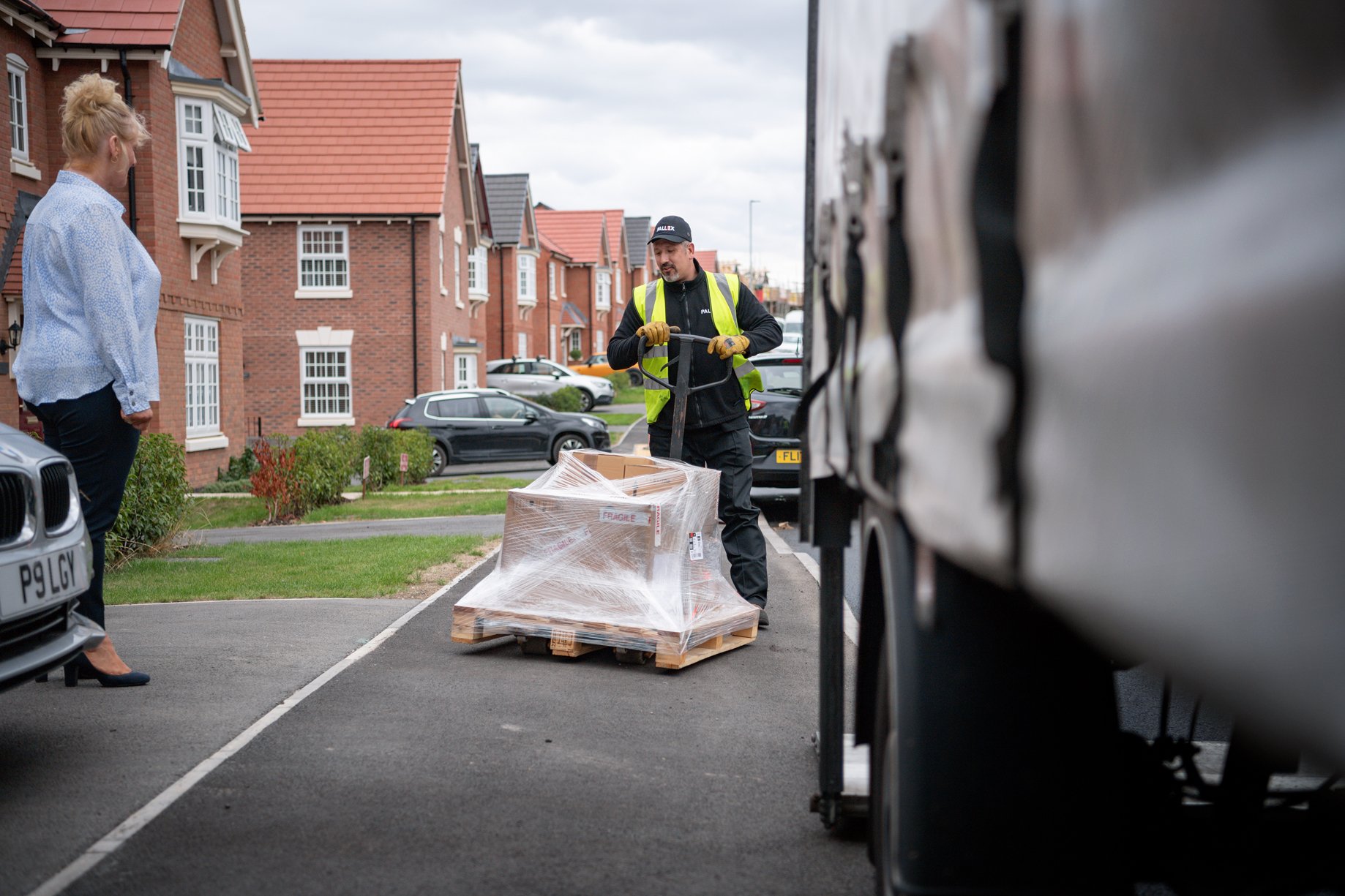 A man in a yellow safety vest is unloading a pallet wrapped in plastic from a delivery truck, while a woman in business attire watches nearby in a residential neighborhood with red brick houses and parked cars.