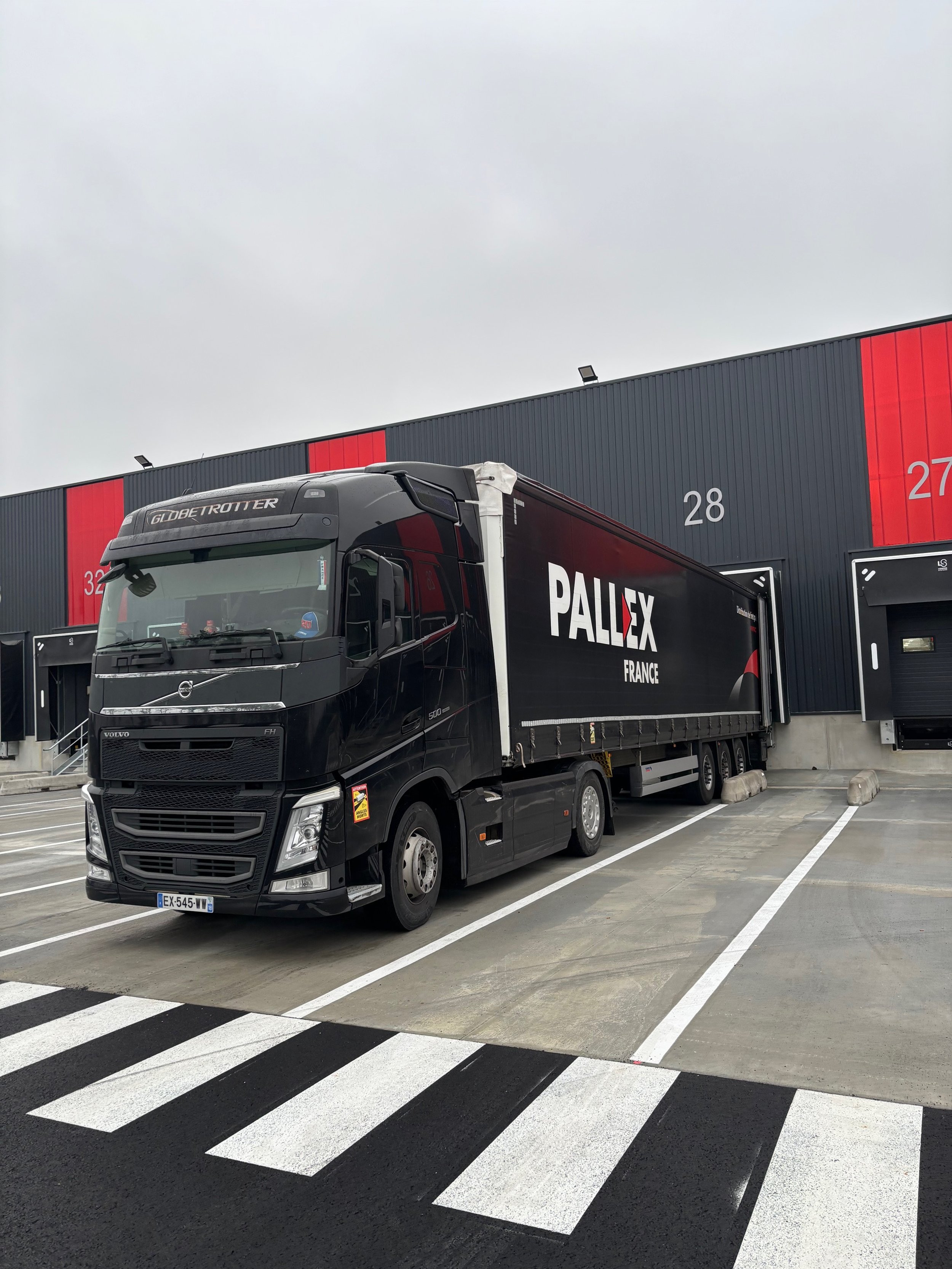 A black Volvo GLOBETROTTER truck parked at a loading dock with the number 28 on a dark-colored warehouse wall.