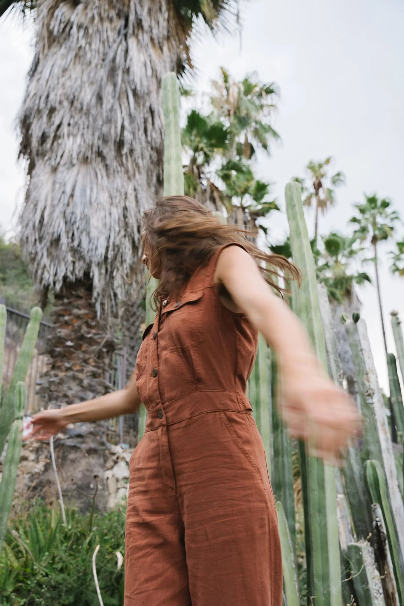 A woman in a rust-colored jumpsuit is dancing or moving among tall cactus plants with a large palm tree in the background.