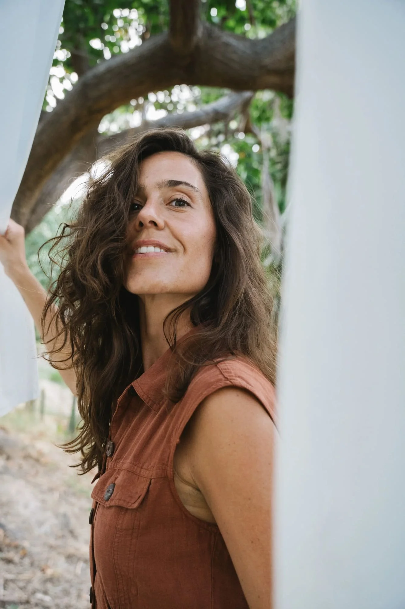 A woman with long wavy brown hair and freckles smiles while standing outdoors near a white wall with a tree in the background.