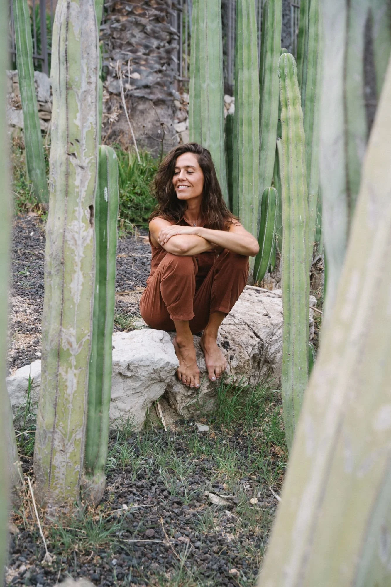 A woman with long brown hair sitting on a large white rock among tall green cacti, smiling and looking to her left.