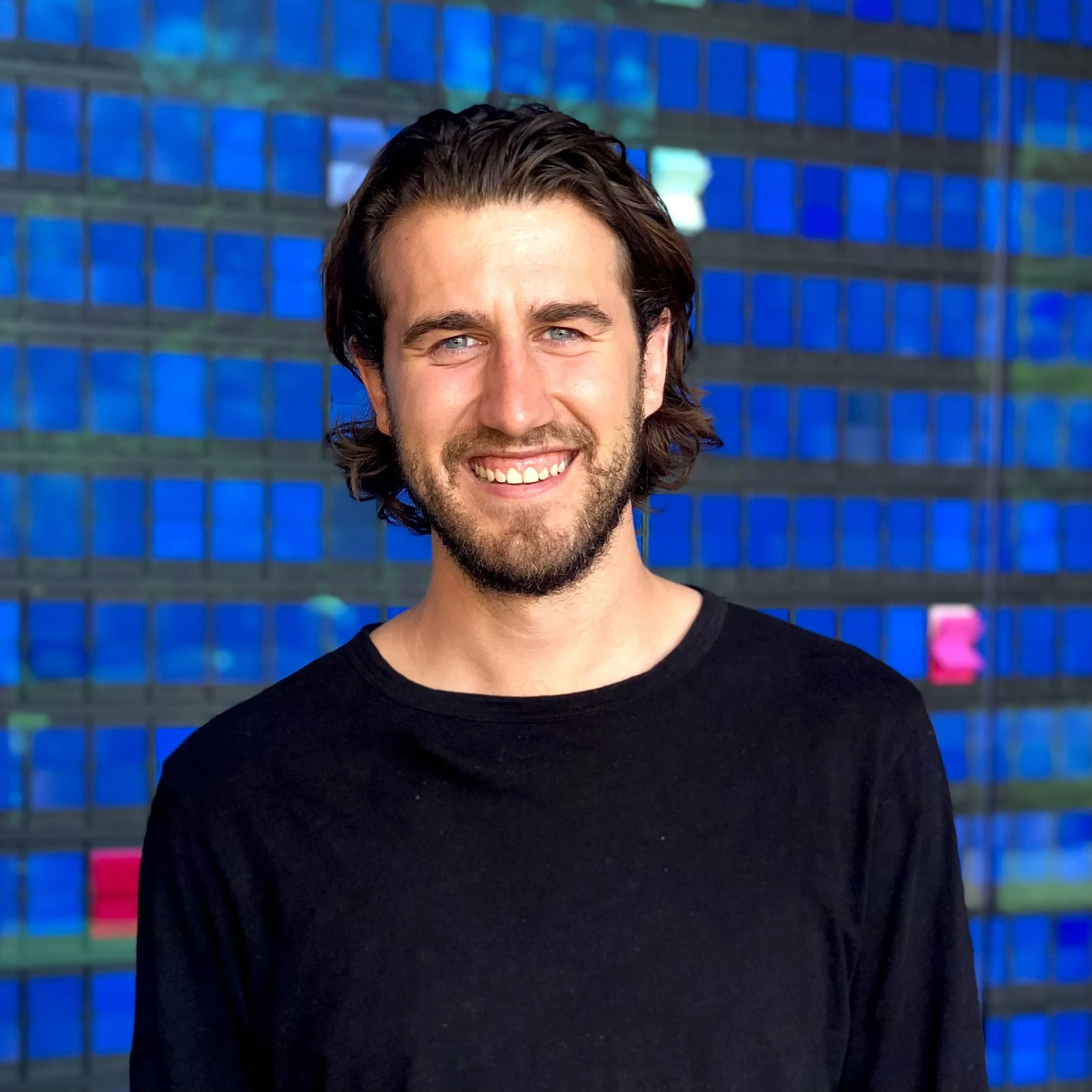 A young man with long dark hair, a beard, and blue eyes smiling in front of a building with a blue glass facade.