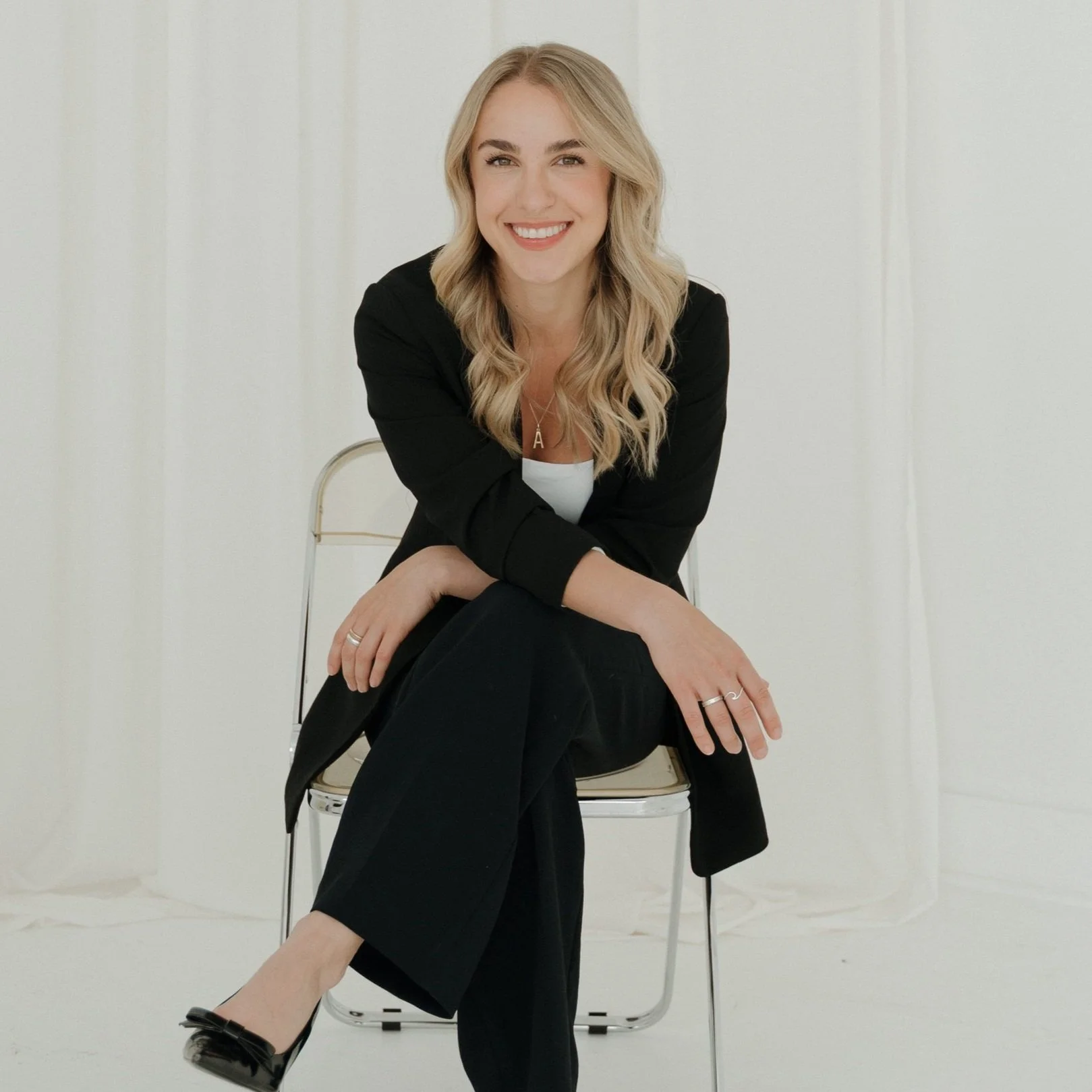 Smiling woman with blonde hair sitting on a chair against a plain white background, wearing a black blazer and black pants.