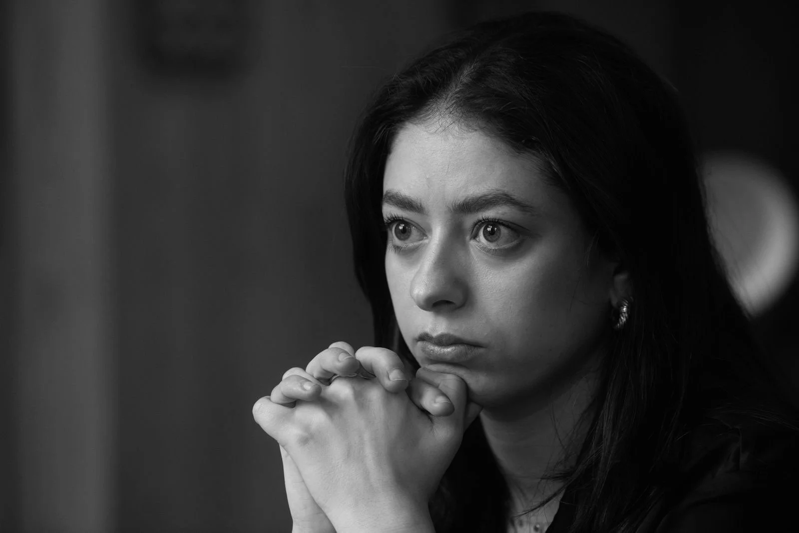 Zwarte en witte portretfoto van een jonge vrouw met lange donkere haar, haar handen onder haar kin geklopt, diep nadenkend of geconcentreerd.