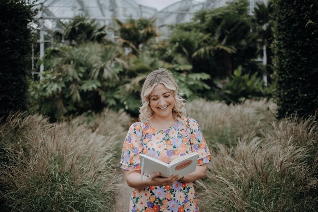 A smiling woman who is a wedding celebrant wearing a colourful floral dress, holding a ceremony script and standing in a lush greenhouse or botanical garden with tropical plants around her.