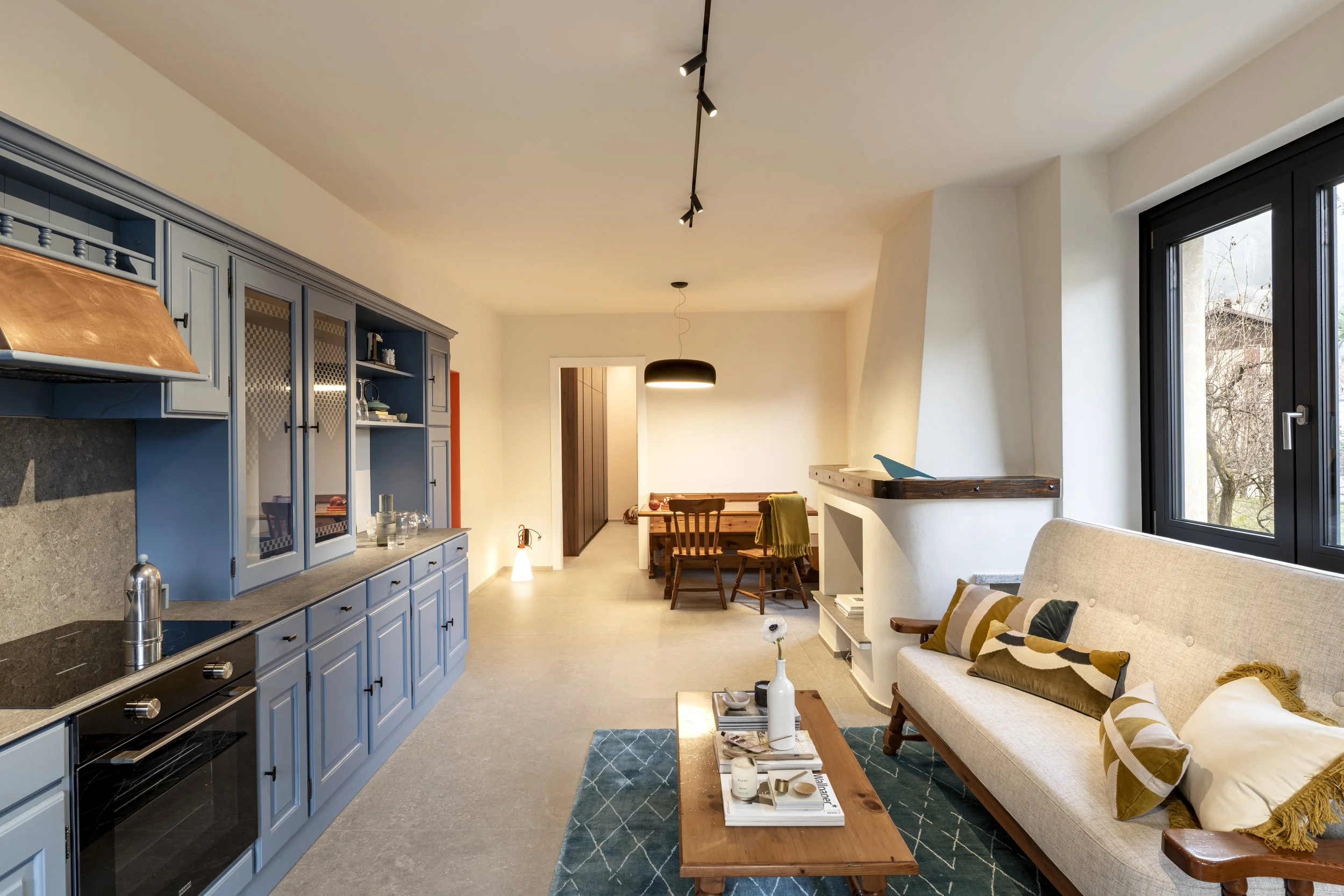 Interno soggiorno con cucina a sinistra, mobili azzurri, piano in pietra, tazza e lavello, e zona pranzo al centro con tavolo in legno e sedie in legno. A destra, un divano beige con cuscini, una finestra grande con vista sugli alberi, e una lampada sospesa sopra il tavolo. Il pavimento è in piastrelle e le pareti sono bianche.