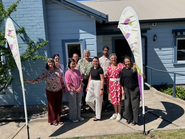 Four women standing in front of a house with a banner that reads "Reimagining the future" and "WITH NRT" on a sidewalk surrounded by green bushes and grass.