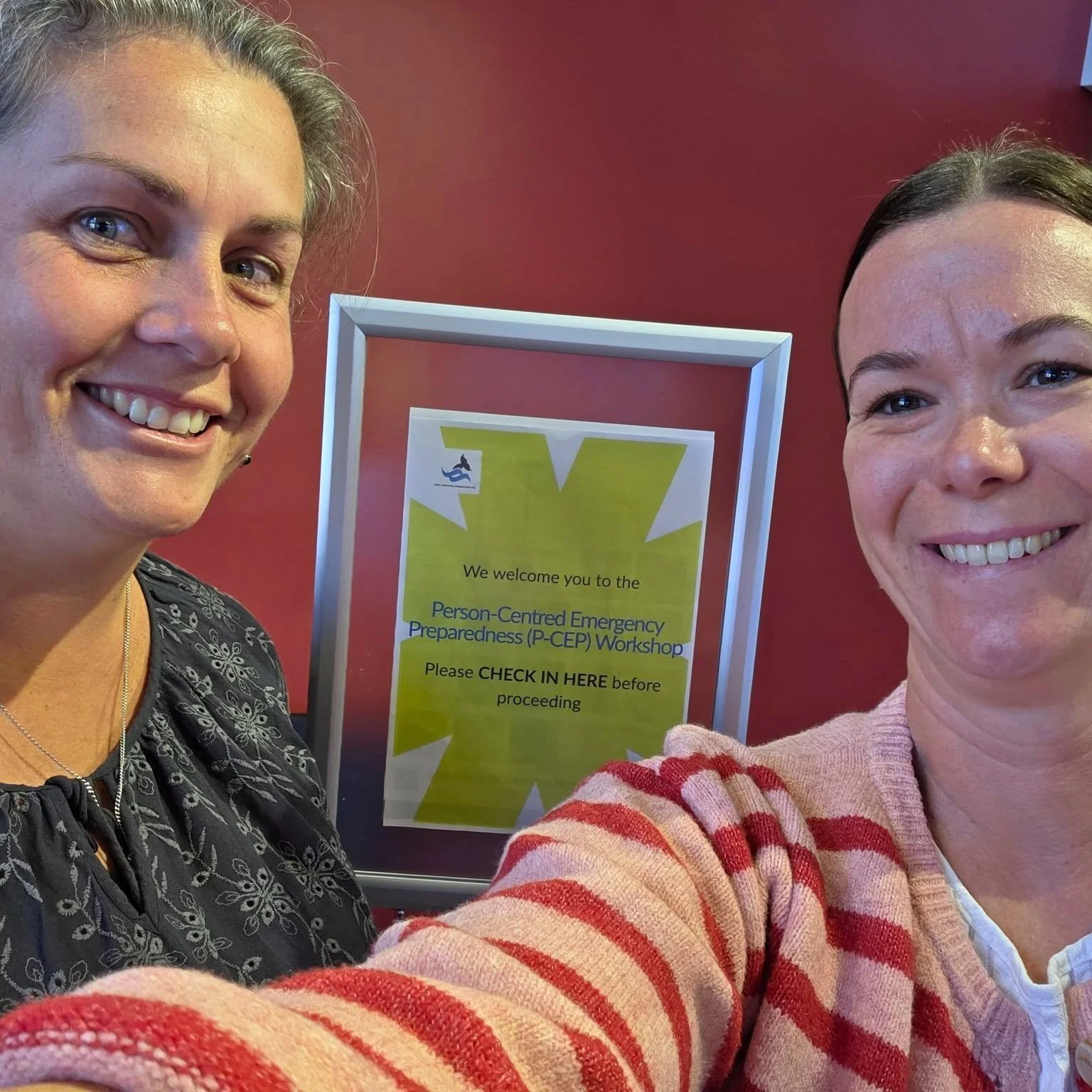 A selfie of two women smiling at a workshop venue. Behind them is a sign welcoming participants to the “Person-Centred Emergency Preparedness (P-CEP) Workshop