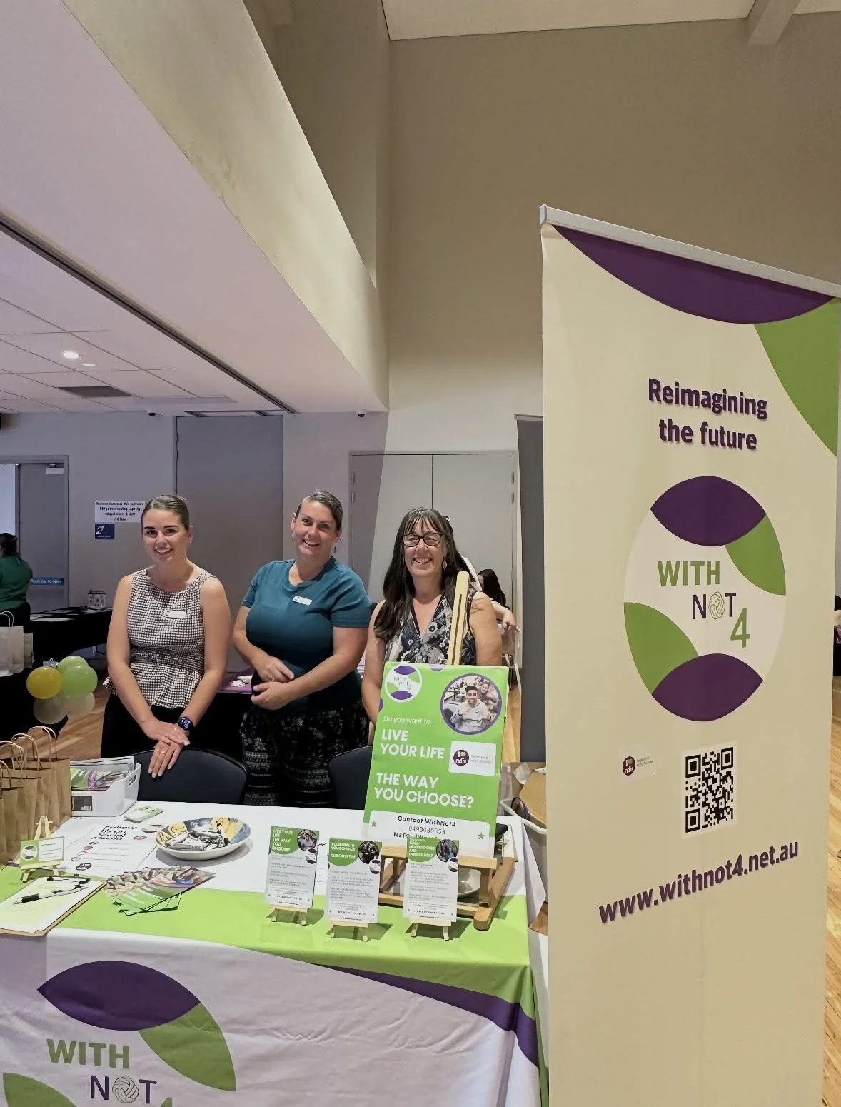 First image/ Three team members smiling behind a branded expo table with brochures, flyers and a vertical banner reading "reimagining the future - With Not 4" in a community hall setting.  Second image/ Two team members standing behind a display tabl