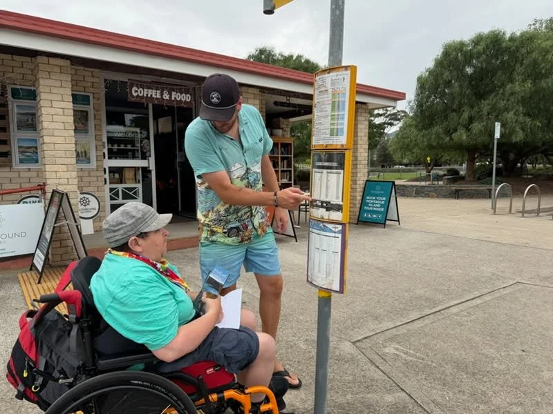 Two people looking at a bus schedule. One seated in a wheelchair, another standing pointing to the schedule at a bus stop
