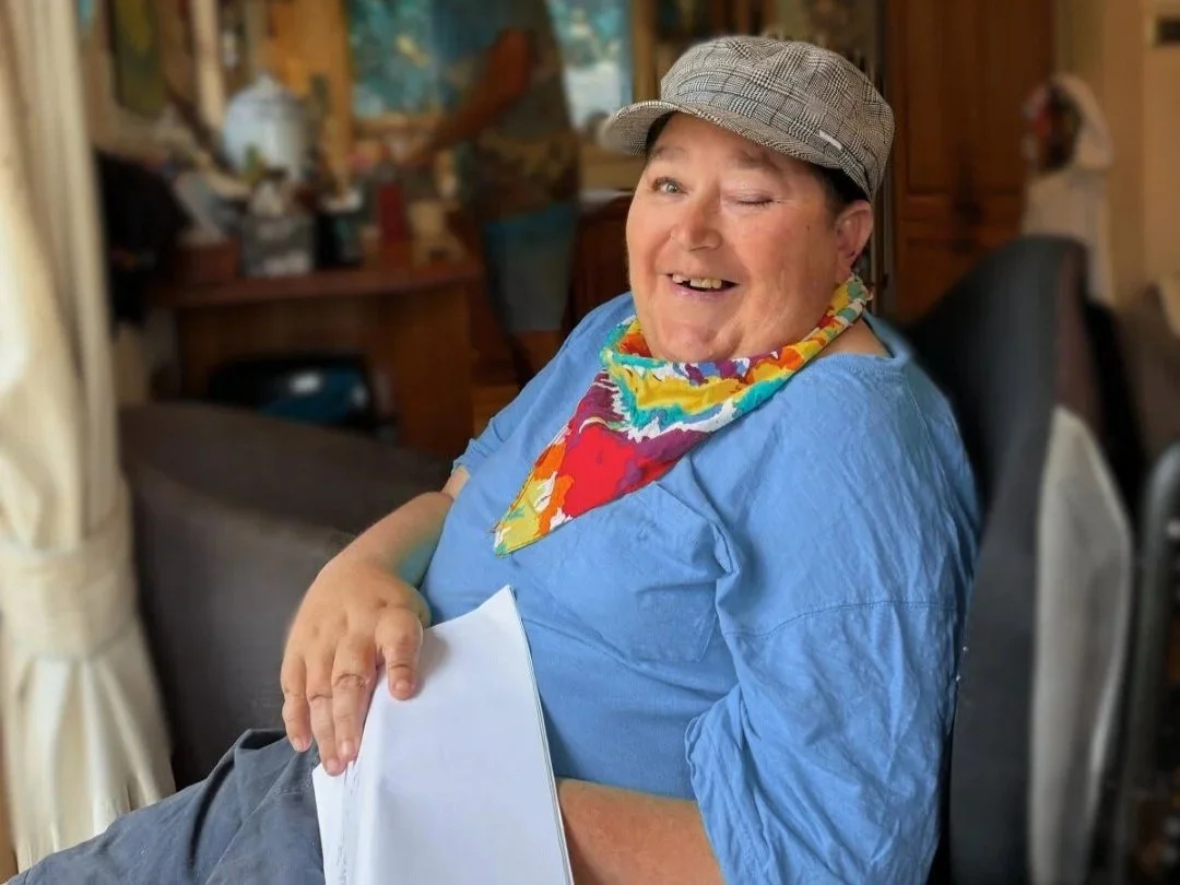 A smiling person wearing a grey cap and colourful scarf sits in a chair indoors, holding papers, with a warm home setting and ILA in the background. Text on the image reads “Invisibility is a skill not a magical super-power” alongside the WithNot4 lo
