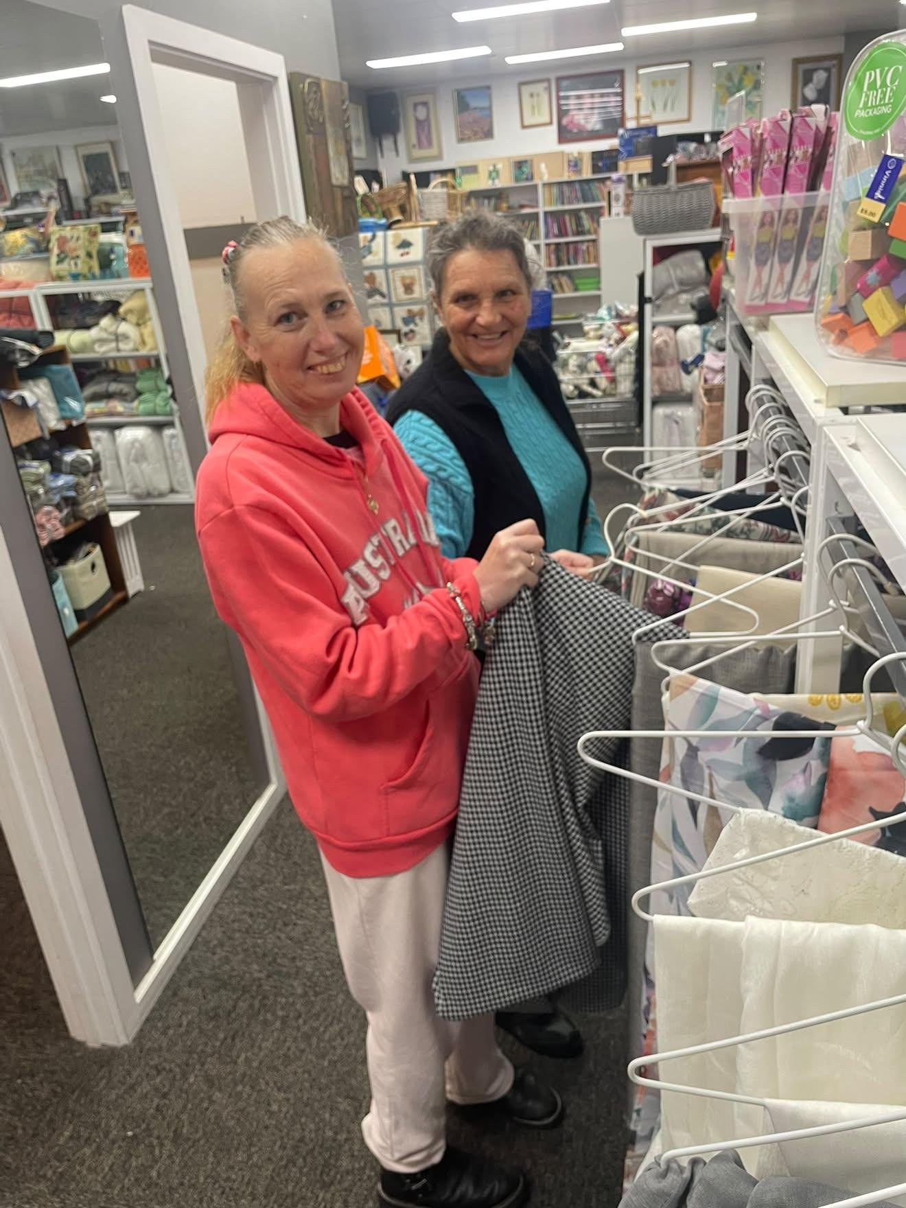  Two Women at an Op Shop smiling and holding fabric hanging on coat hangers. 