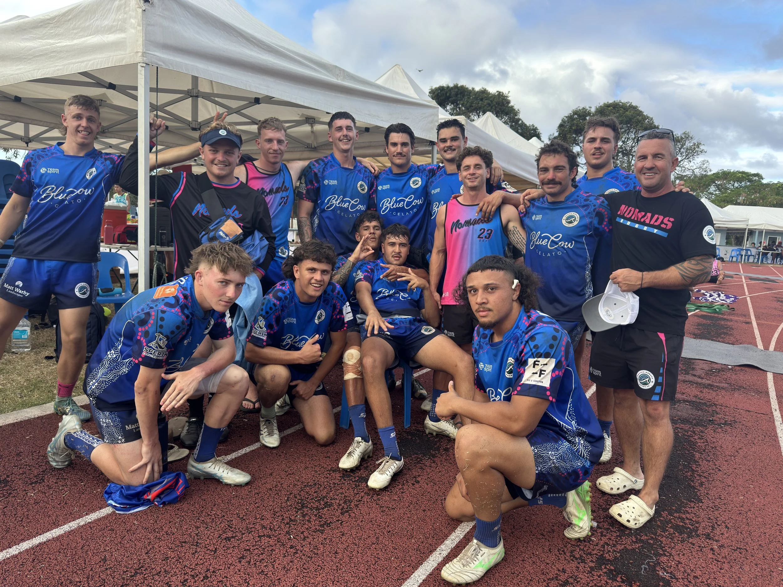 A group of young men and their coach posing on a running track after a sports event. They are wearing blue and pink athletic jerseys, some kneeling and others standing, showing camaraderie and team spirit.