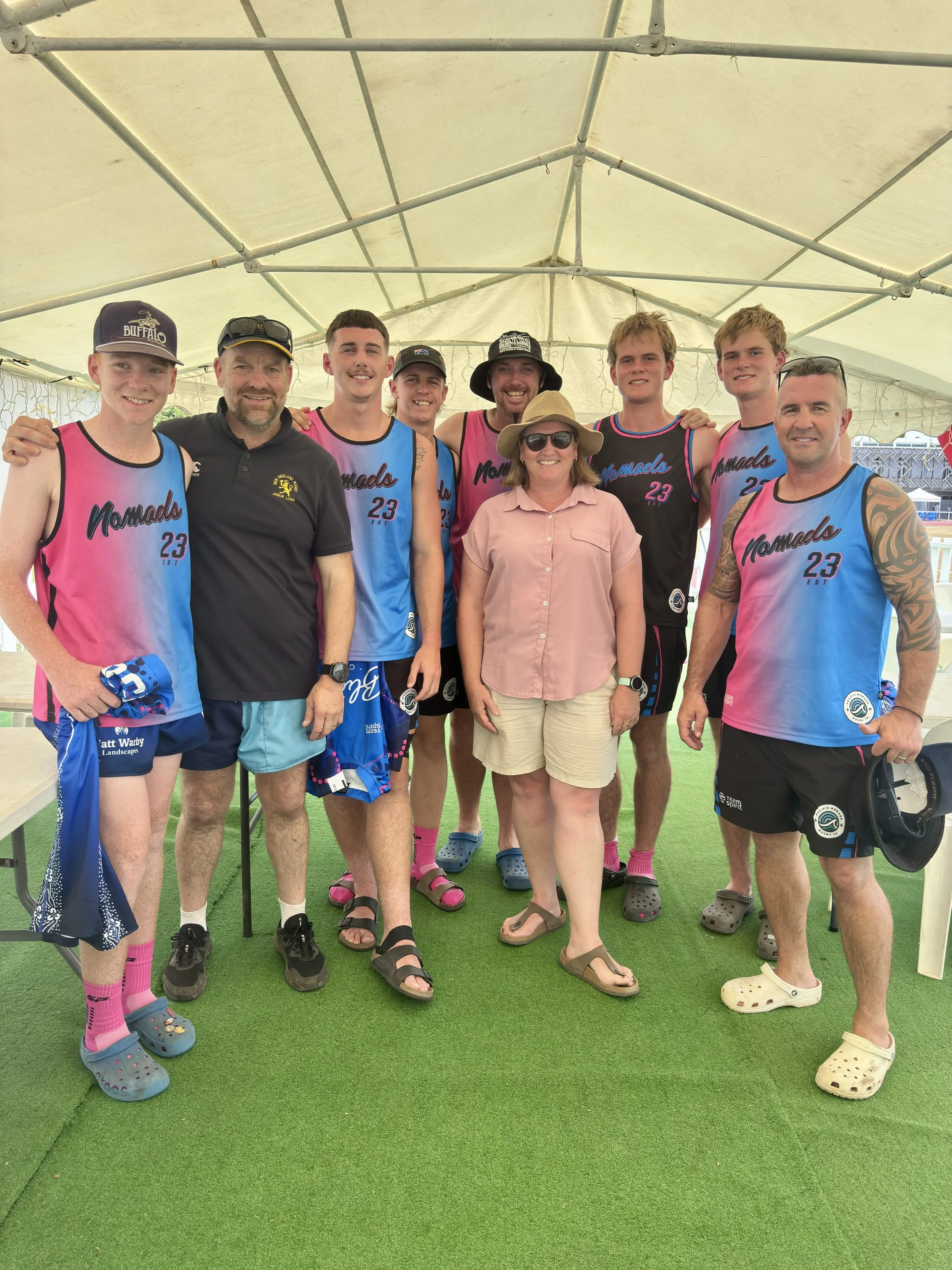 A group of eight people, six young men and two women, posing together inside a white canopy tent, with some wearing matching sports jerseys that read 'Nomads 23', representing a sports team. They are standing on artificial turf, smiling at the camera