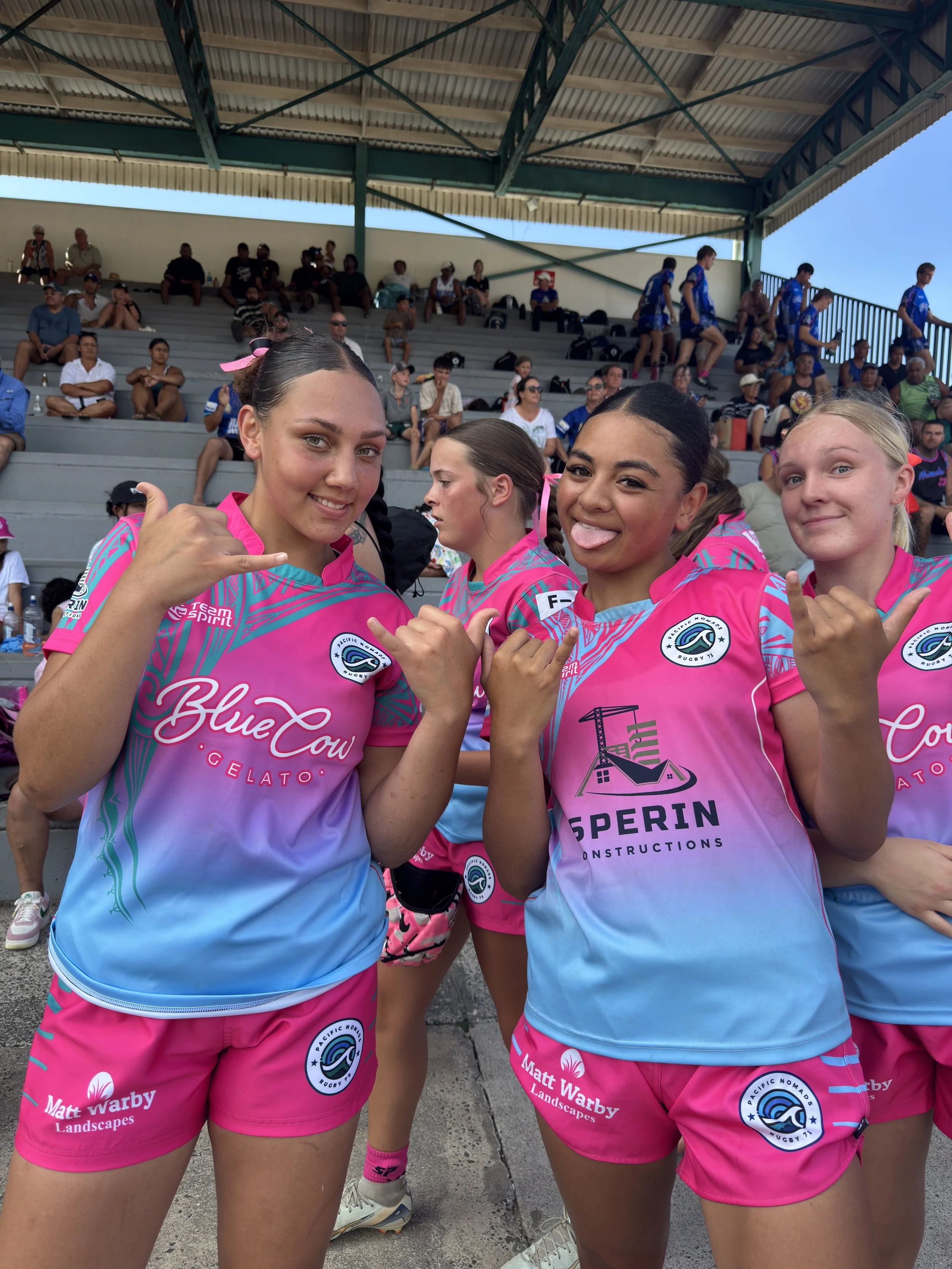 Group of female rugby players in pink and blue uniforms posing outdoors with spectators in the background.