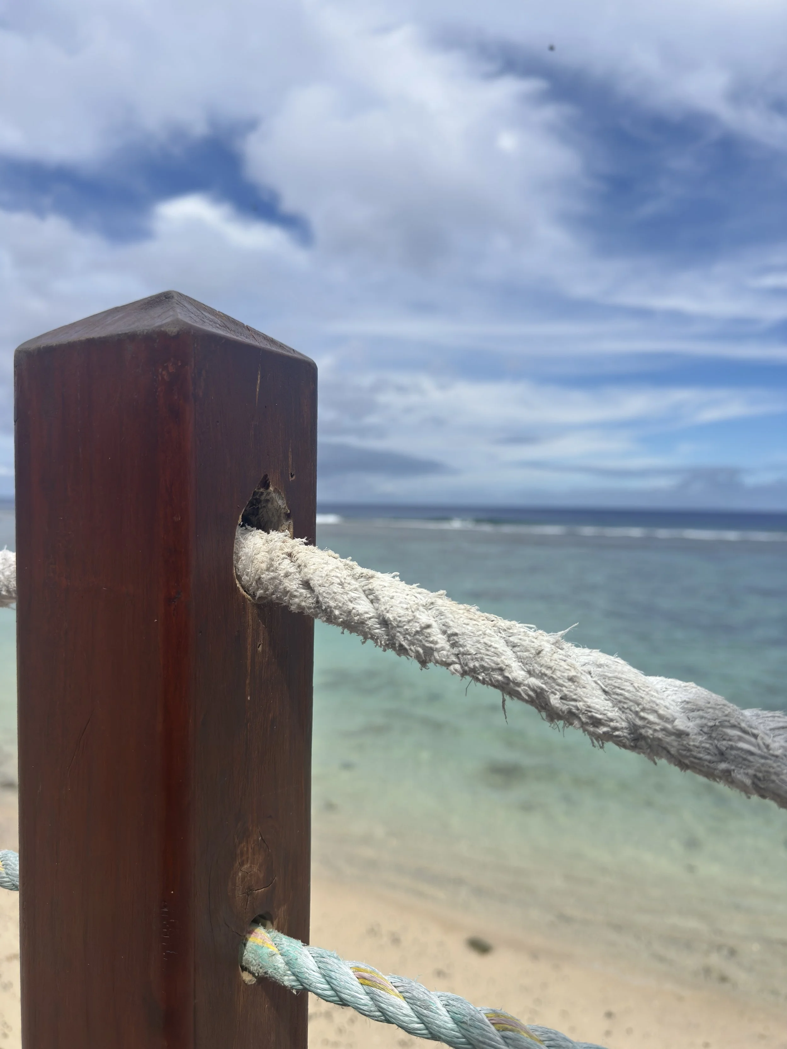 Close-up of a wooden post with frayed rope tied through a hole, overlooking a beach with sand, water, and cloudy sky.