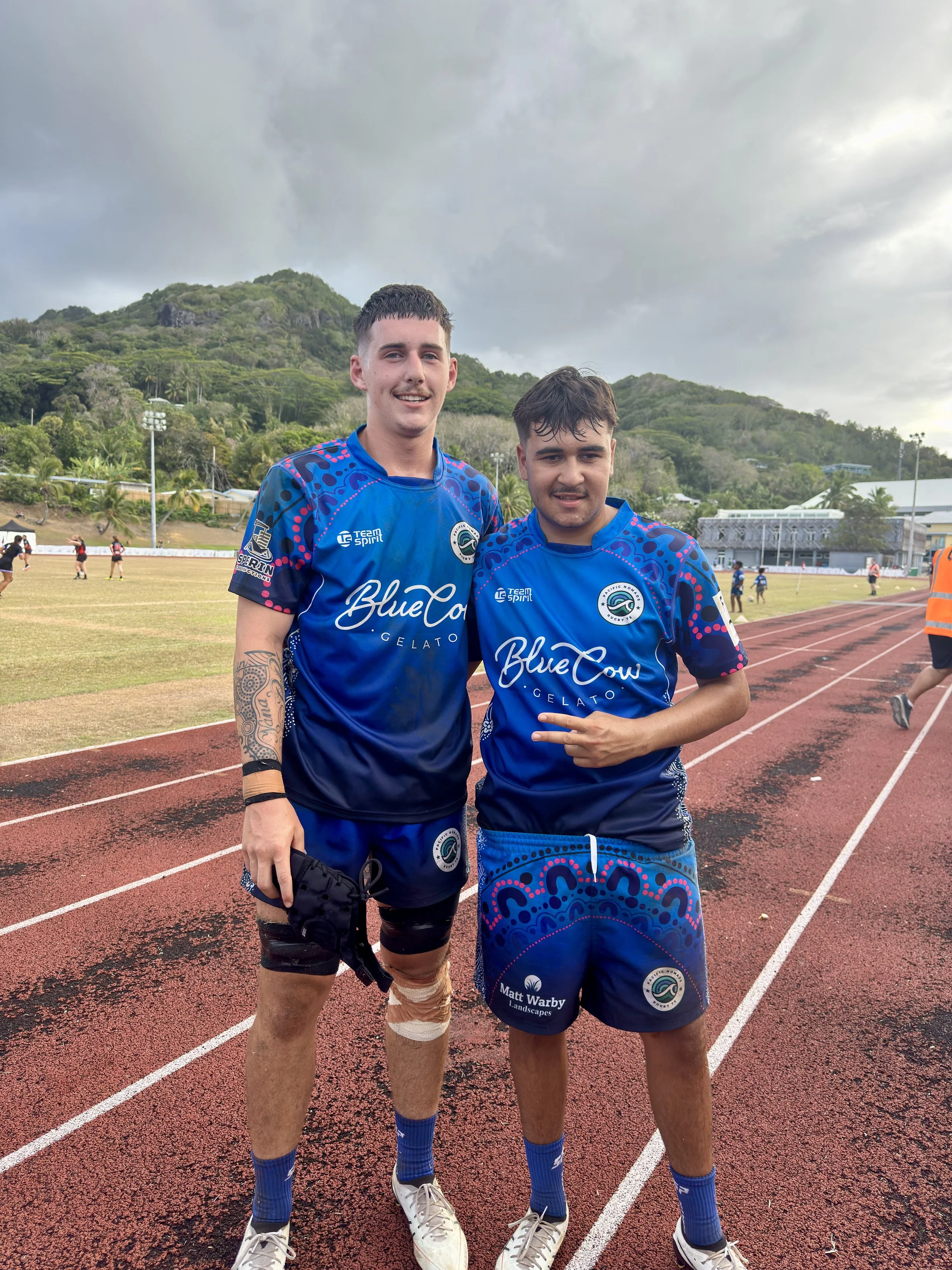 Two young men in blue sports uniforms standing on a running track, with a grassy field and hills in the background. One is holding a hat, and they are making a peace sign. Other players and a person in an orange vest are visible in the background.
