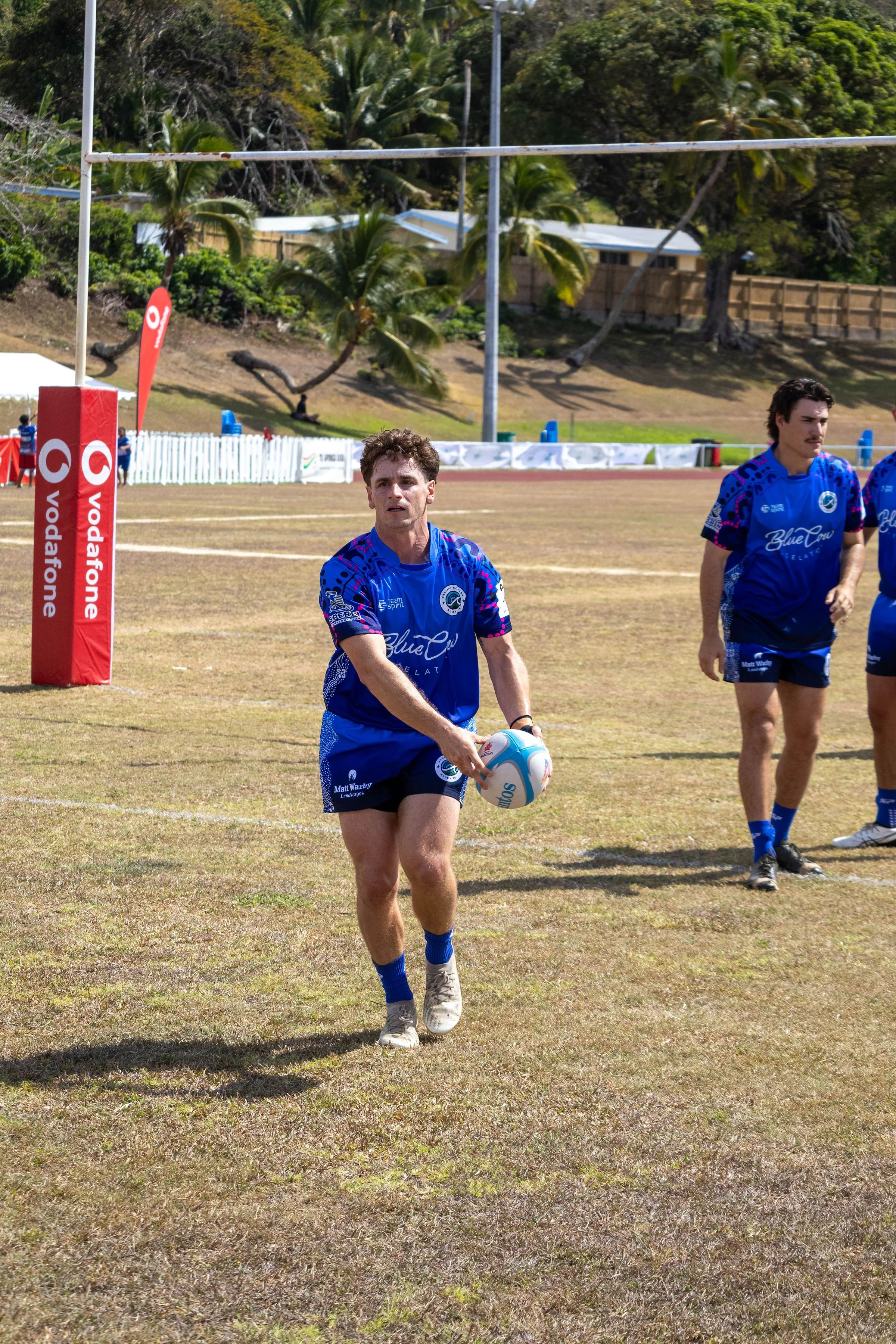 A rugby player in a blue jersey holding a rugby ball on a grassy field, with other players nearby, palm trees, and a stadium in the background.
