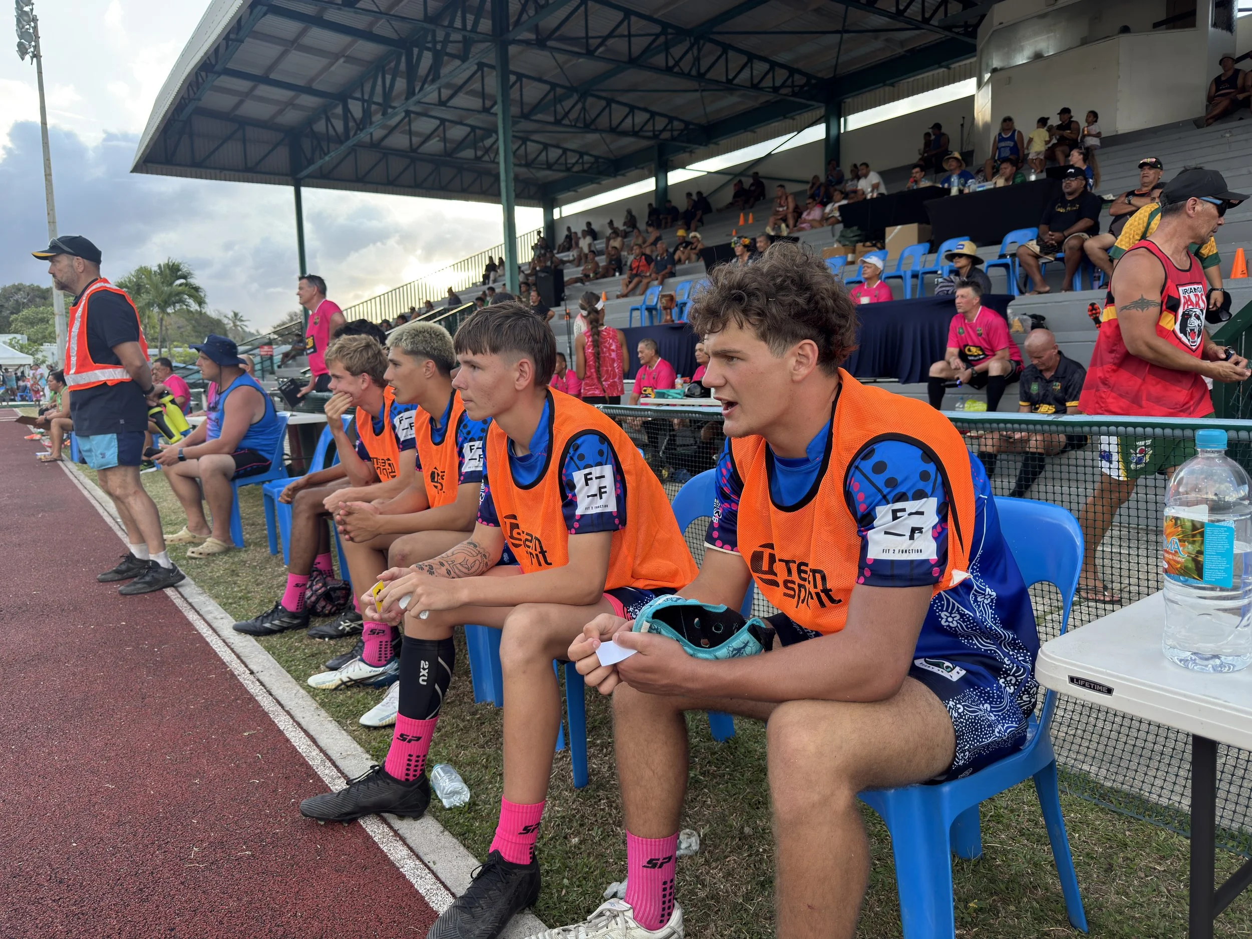 Young athletes sitting on blue chairs by the track during a sporting event, with spectators on the stands behind them.