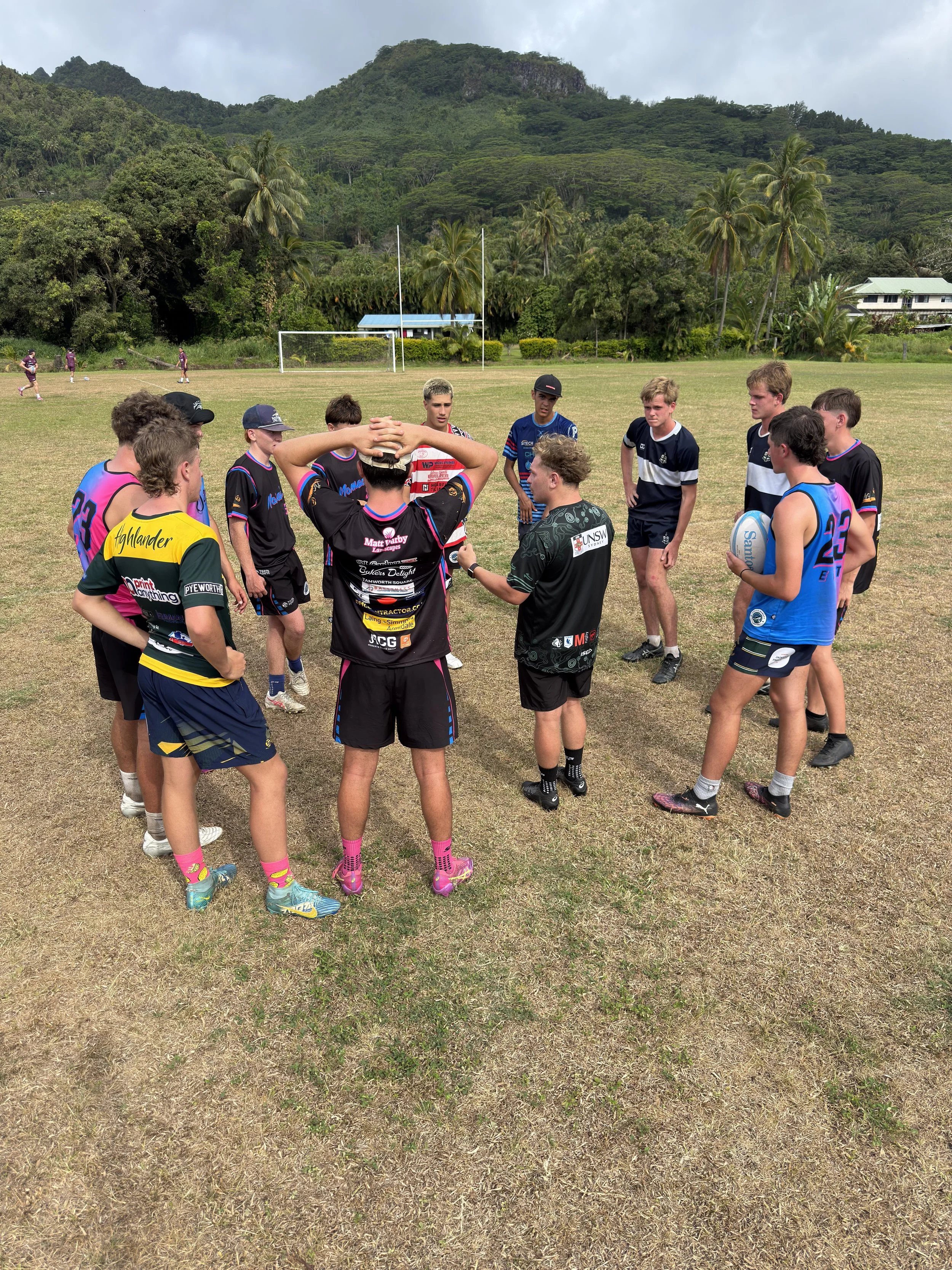A group of young rugby players gathered in a circle on a grassy field, listening to their coach during a practice session, with mountains, palm trees, and a small soccer goal in the background.