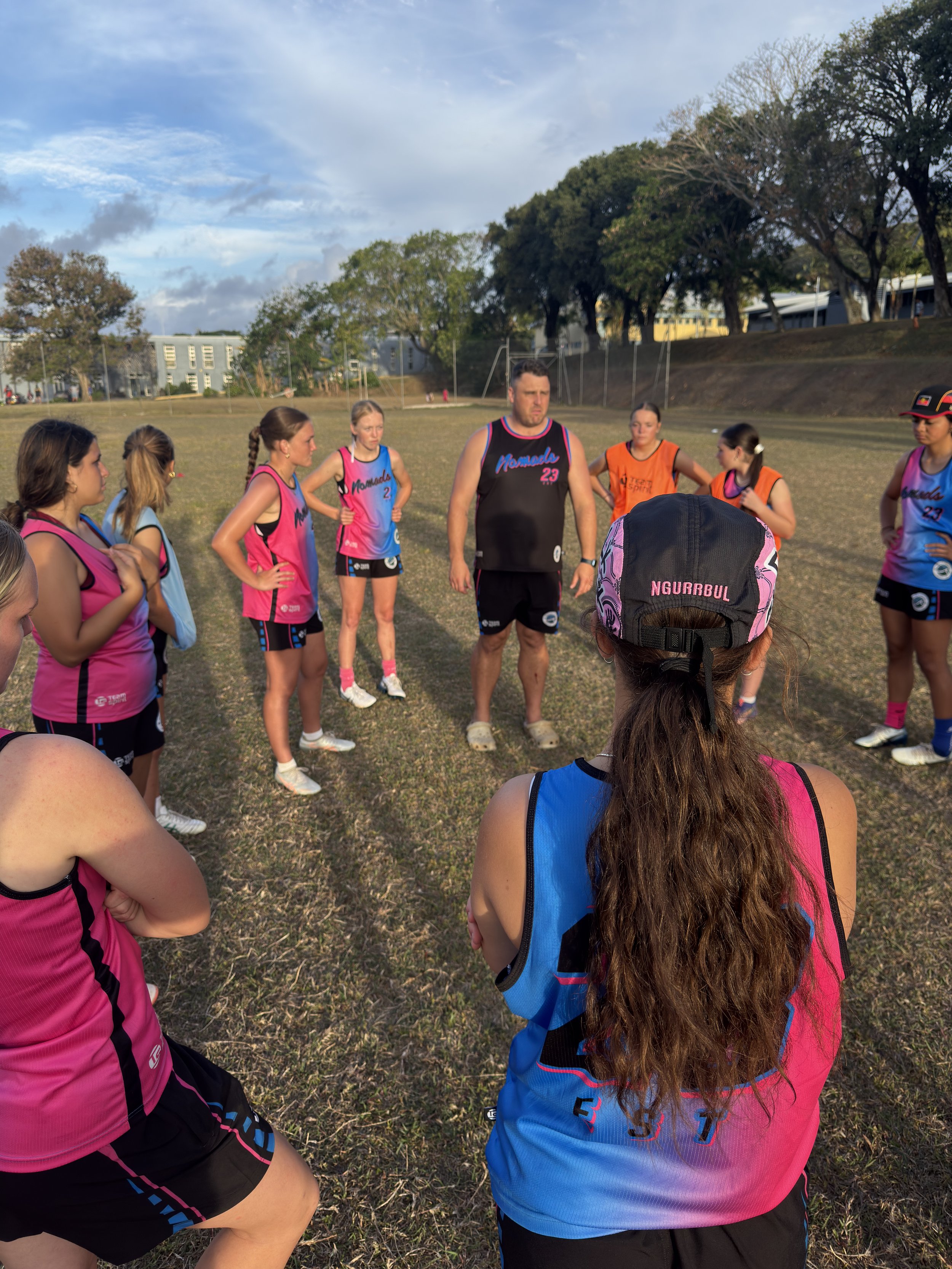 A group of women and a man in athletic gear gathered on a grassy field outdoors, listening to a woman with long hair in a blue and pink sports jersey, during what appears to be a team huddle or training session. The scene takes place under a partly c