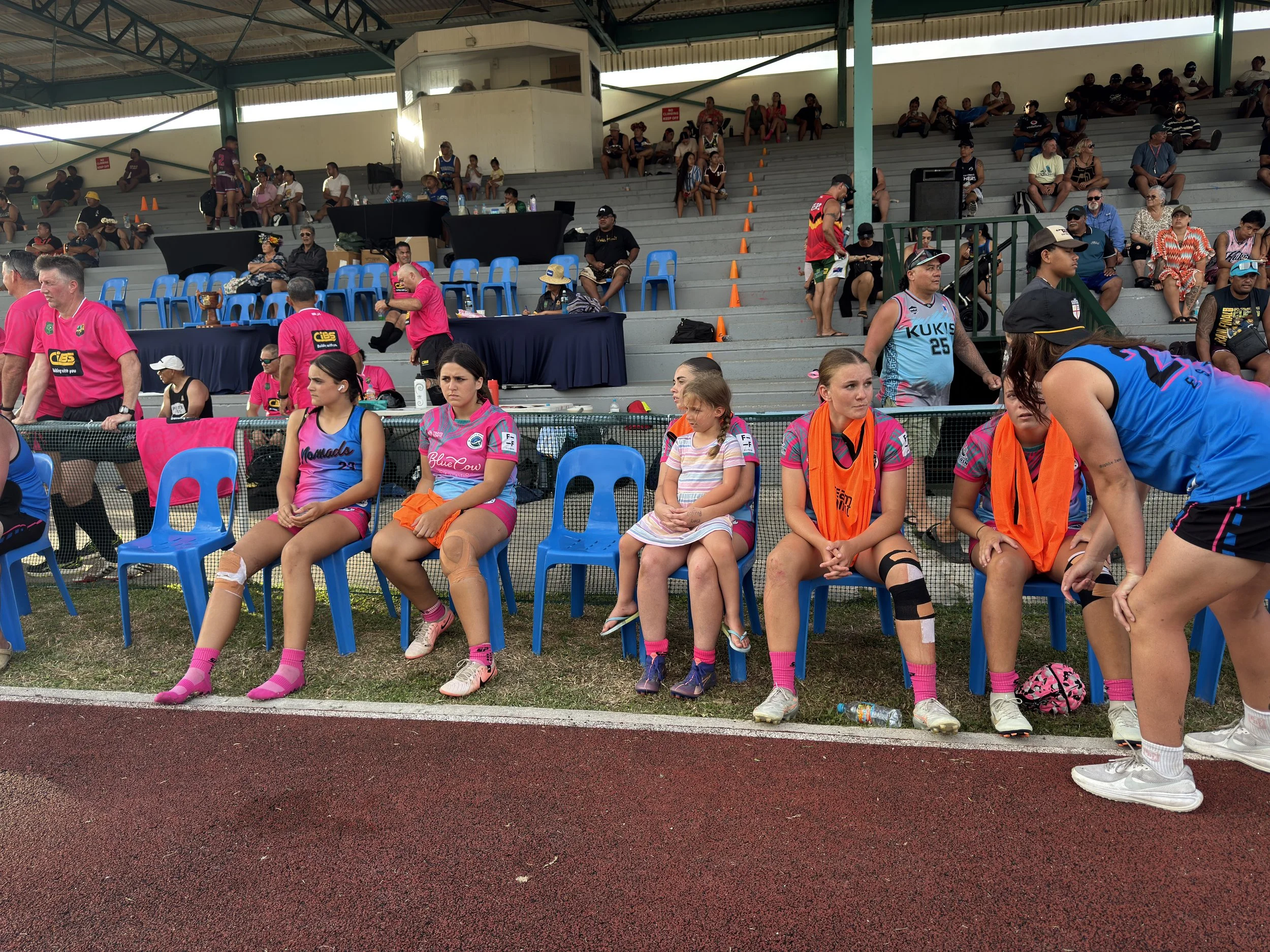 Athletic women sitting on blue chairs at a sporting event, with spectators in the background. Some women are dressed in sports uniforms, with pink socks and knee braces, indicating a team or game setting.