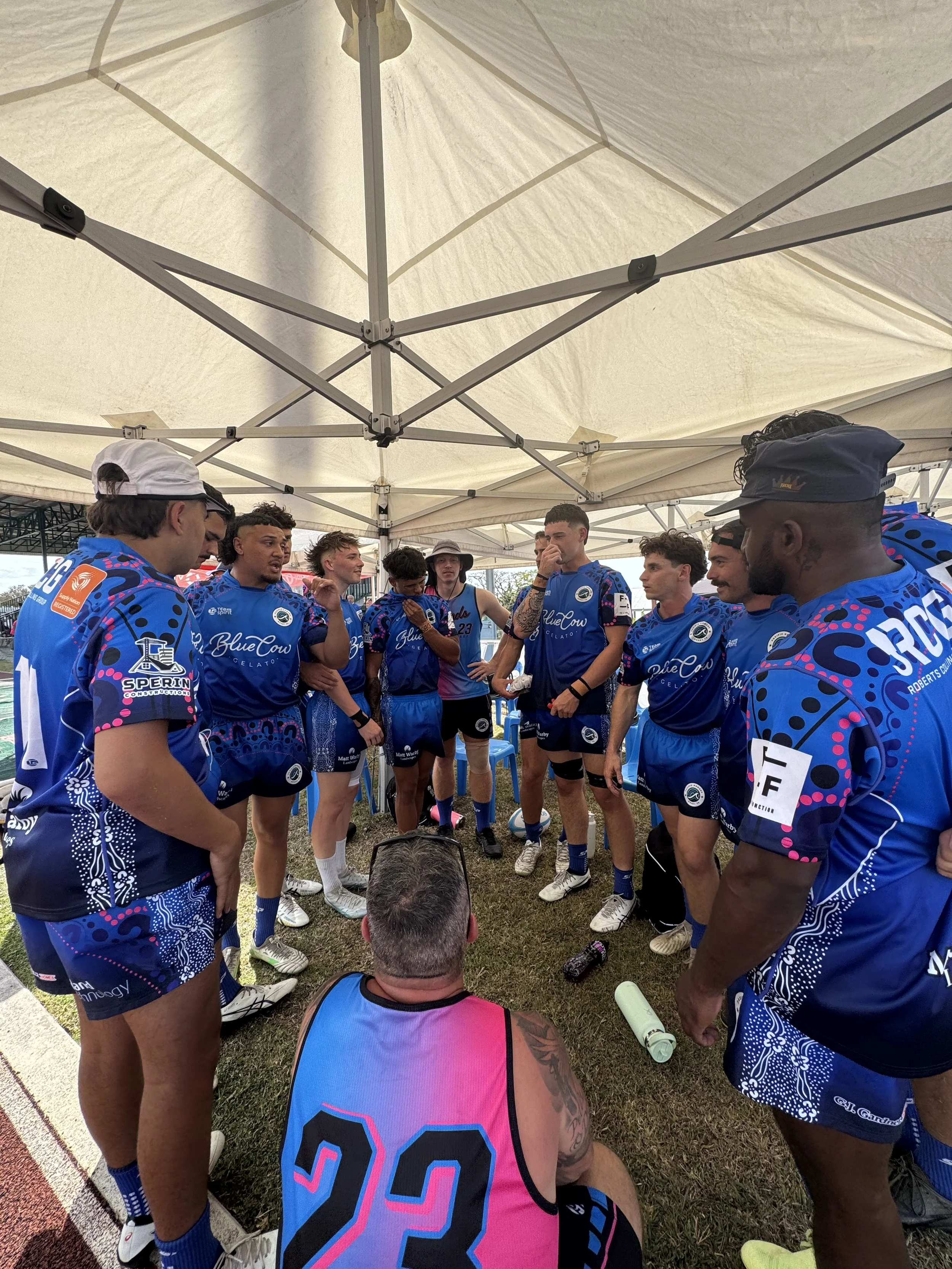 A group of male athletes in blue sports uniforms gathered in a circle under a white canopy, listening to a person in a pink and blue jersey with the number 22 on the back. They appear to be having a team meeting or strategy discussion.
