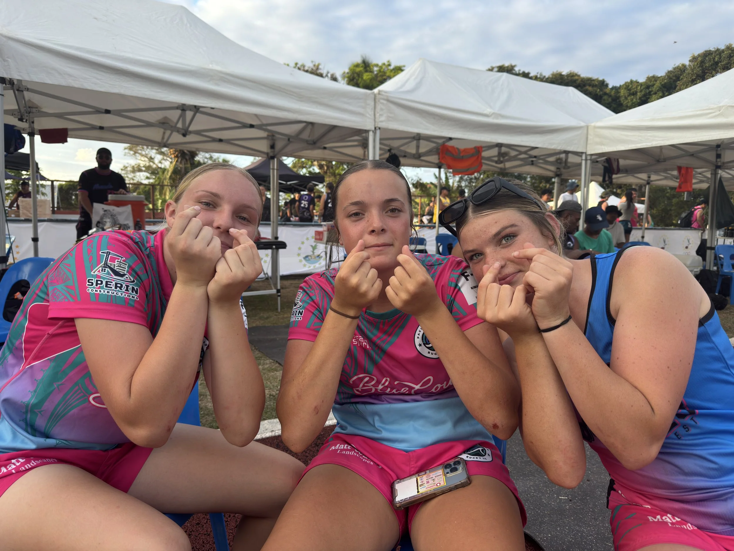 Three young women sitting outdoors under white tents, making finger heart gestures, at an event with spectators and vendors in the background.
