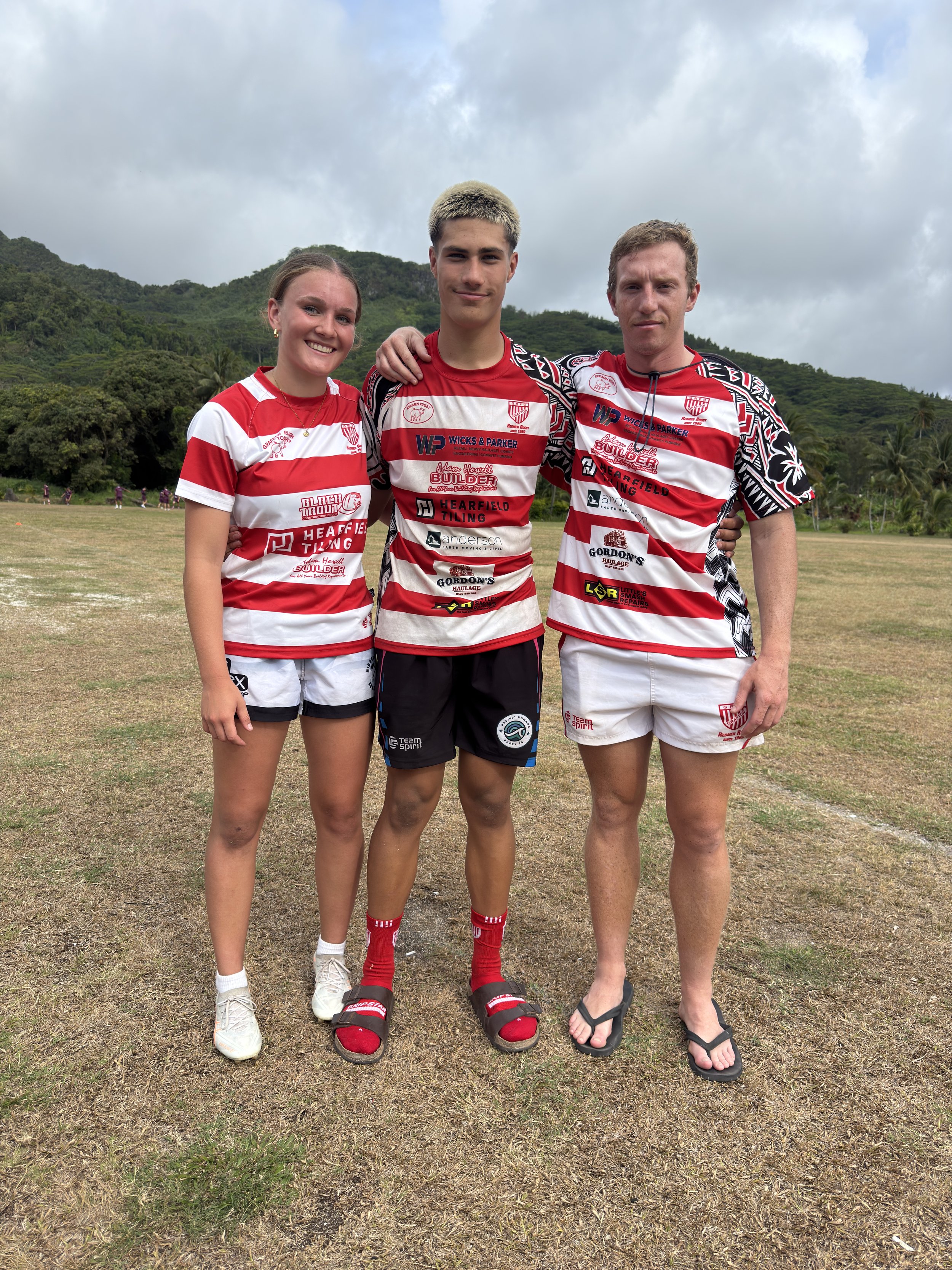 Three young people standing outdoors on a sports field, wearing red and white striped sports uniforms, smiling at the camera.