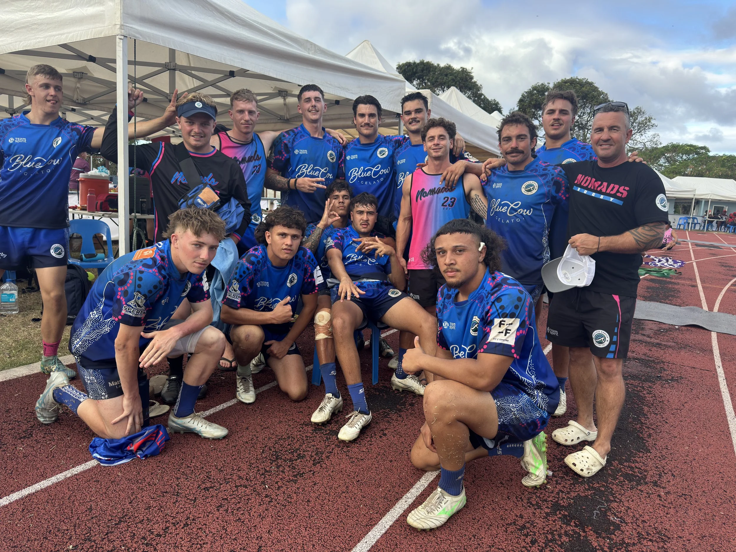 A group of young male athletes and their coach pose for a photo on a running track under a white canopy tent. The athletes wear blue sports jerseys, some with pink accents, and athletic shoes. The coach wears a black t-shirt with pink text. There are