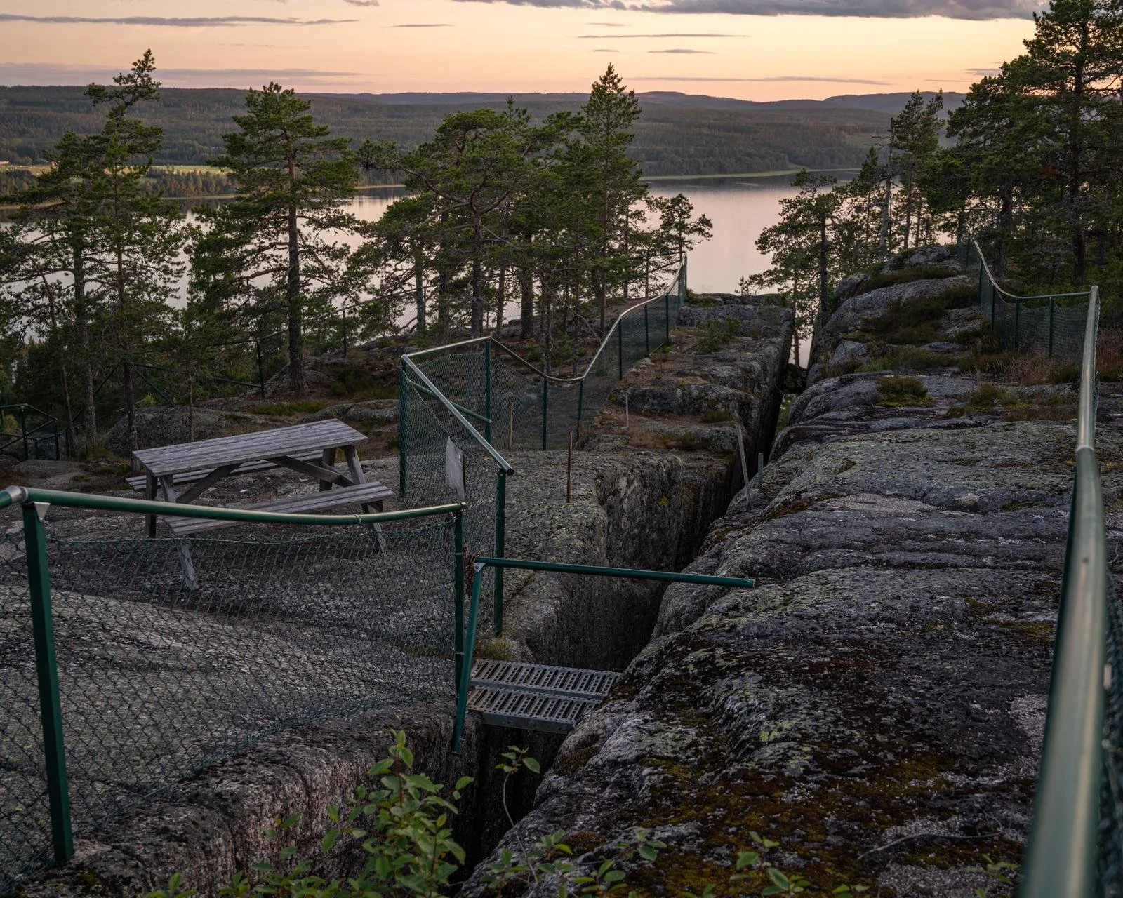 Utsikt över en sjö omgiven av skog, med klippor och ett satträcke, under en dramatisk himmel vid solnedgång.