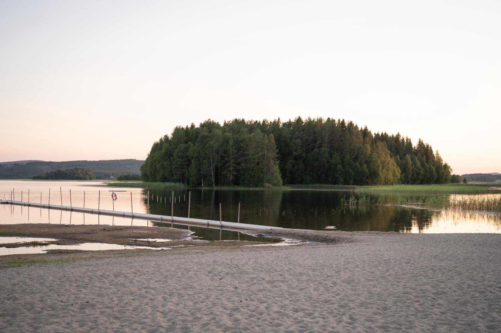 Gröna gräsmattor vid en sjö med en liten brygga och träd i bakgrunden, övervakad av ett molnigt himmel.