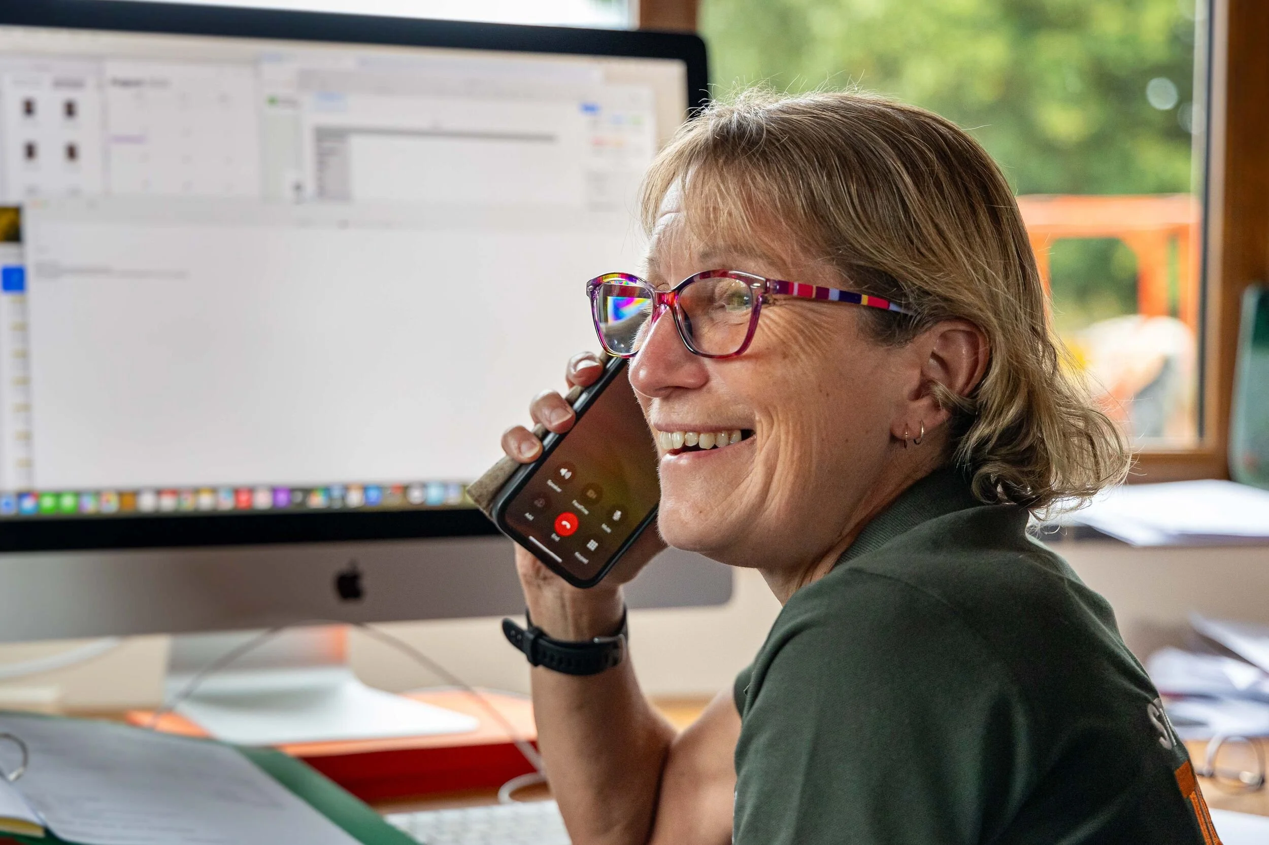 Lady with glasses smiling while talking on a phone to a customer and with a computer screen in the background.