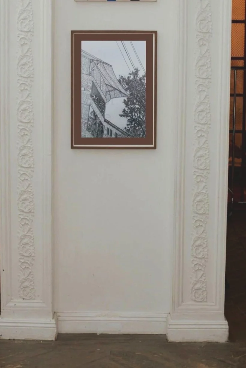 A framed black and white drawing of a building, trees, and power lines on a white wall with ornate molding.