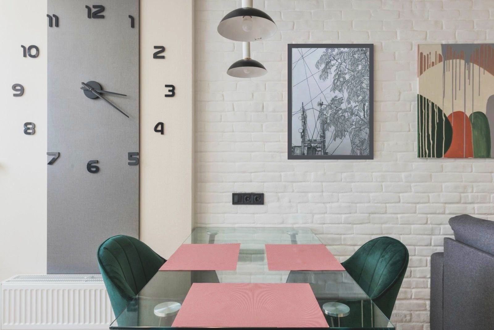 Interior of a dining area with a glass table, three pink placemats, green upholstered chairs, a white brick wall, a large modern wall clock, and framed artwork on the wall.
