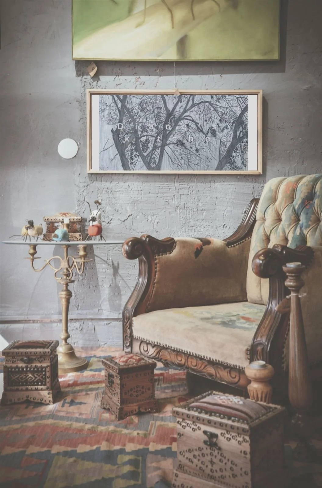 Vintage-inspired cozy seating area with an ornate wooden armchair or sofa, a small round table with decorative items, and a colorful rug with small wooden boxes on the floor. Wall art includes a framed black-and-white picture of trees, and the space has a rustic, textured wall.