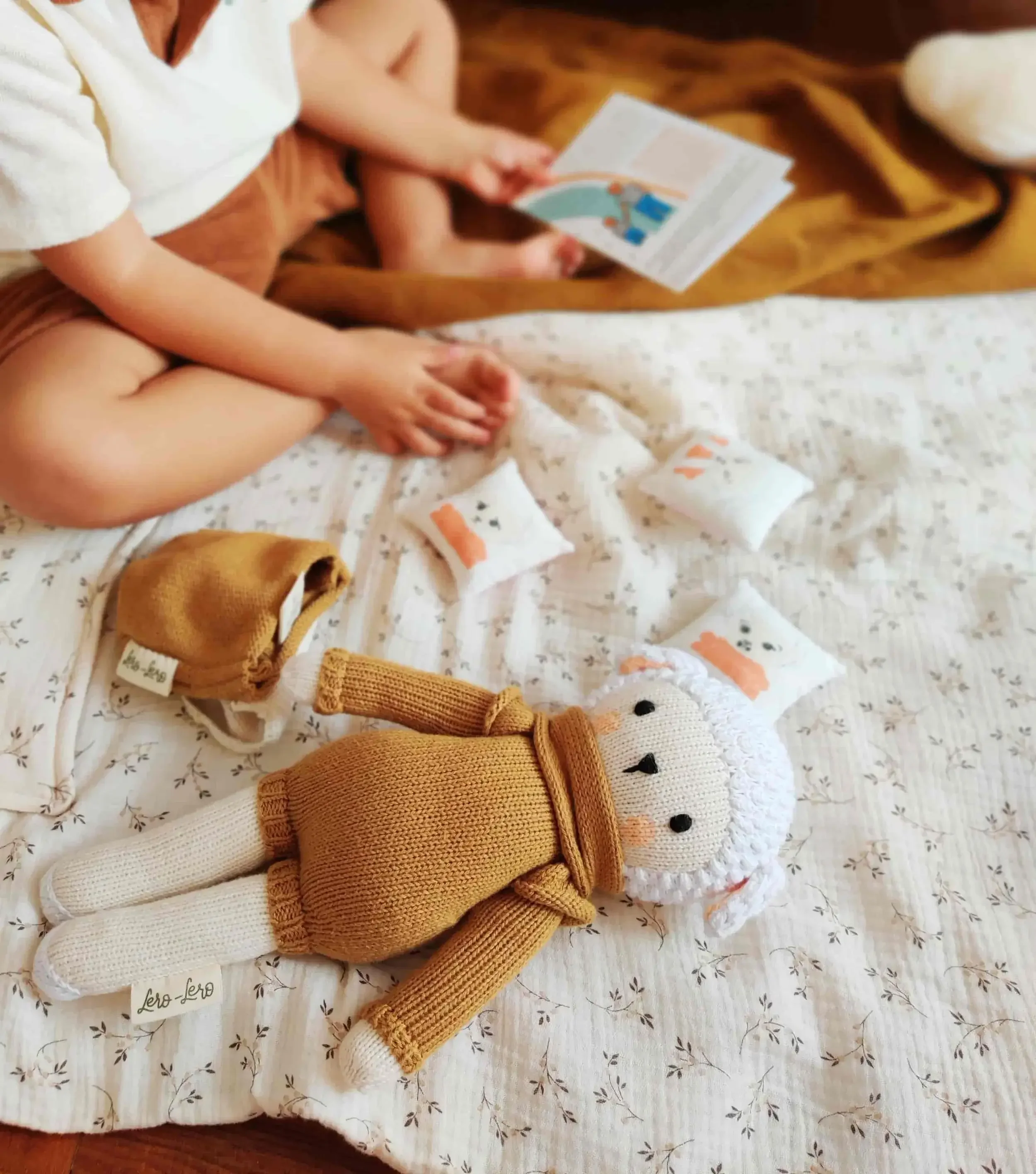 Un enfant assis sur un lit, jouant avec une peluche tricotée en coton bio et plusieurs petits coussins avec des images d'émotions.