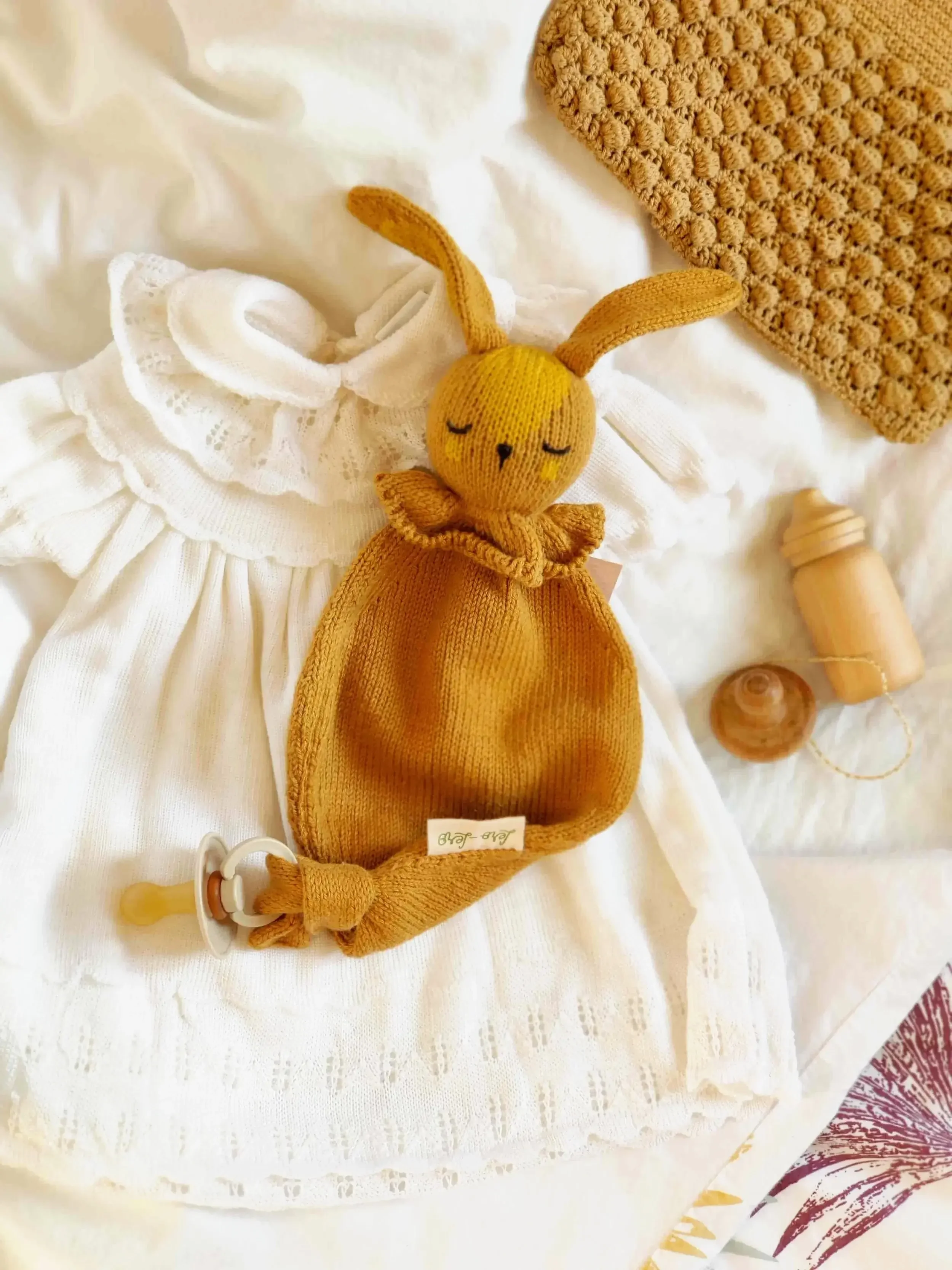 Doudou lapin attache-tétine tricoté artisanalement en coton bio couleur caramel sur un lit avec une tétine et un biberon