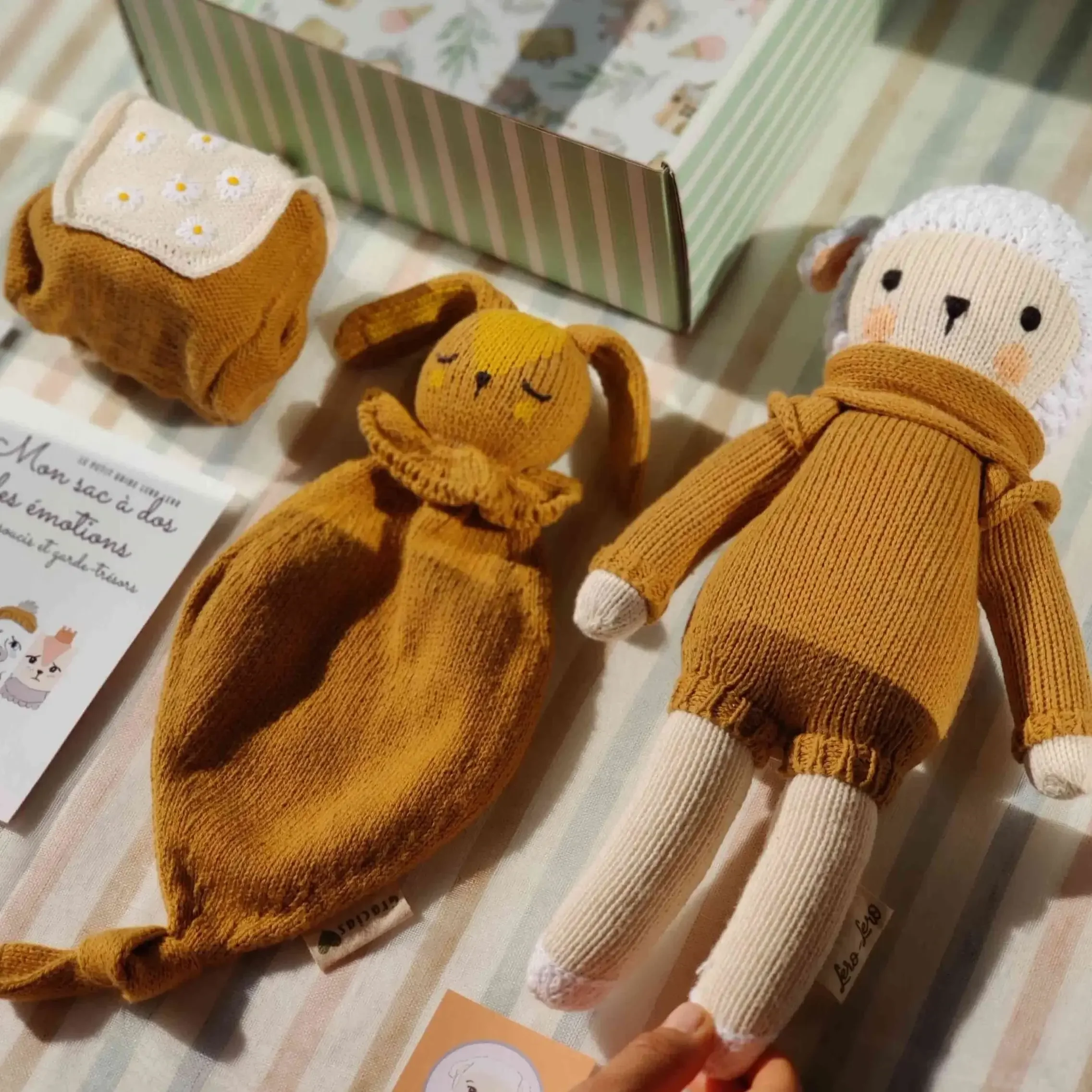 Détail sur la composition du coffret naissance Nino Caramel le Mouton : peluche Nino Caramel, sac à dos des émotions à broderies fleurs et attache-tétine. Tricotés en coton bio.