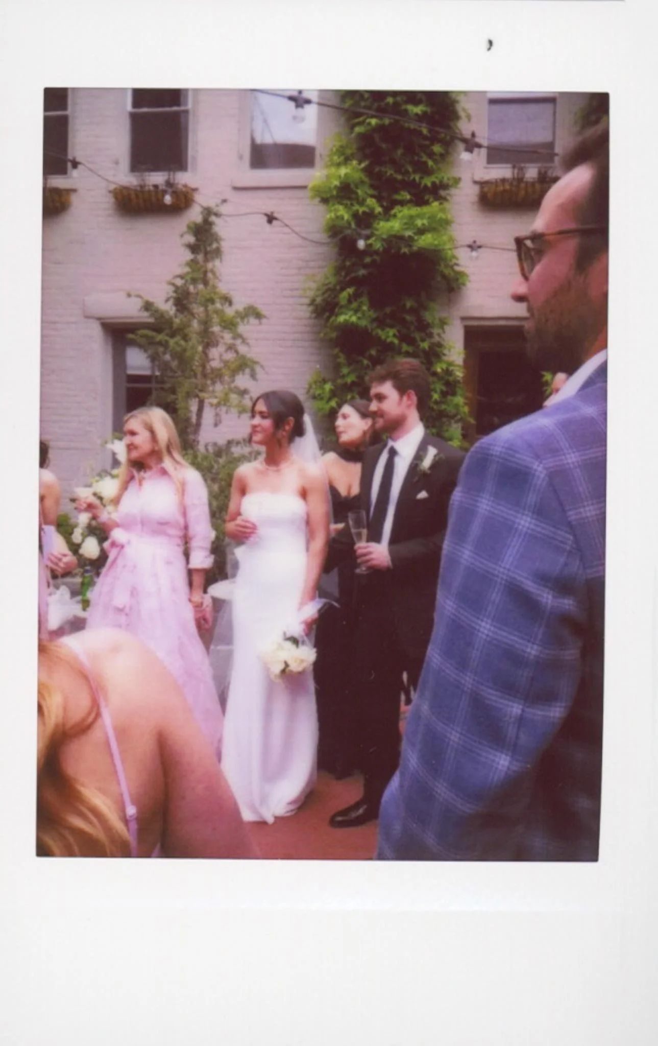 People at a wedding celebration outdoors, with women in dresses and men in suits, standing in front of a building with greenery and string lights.