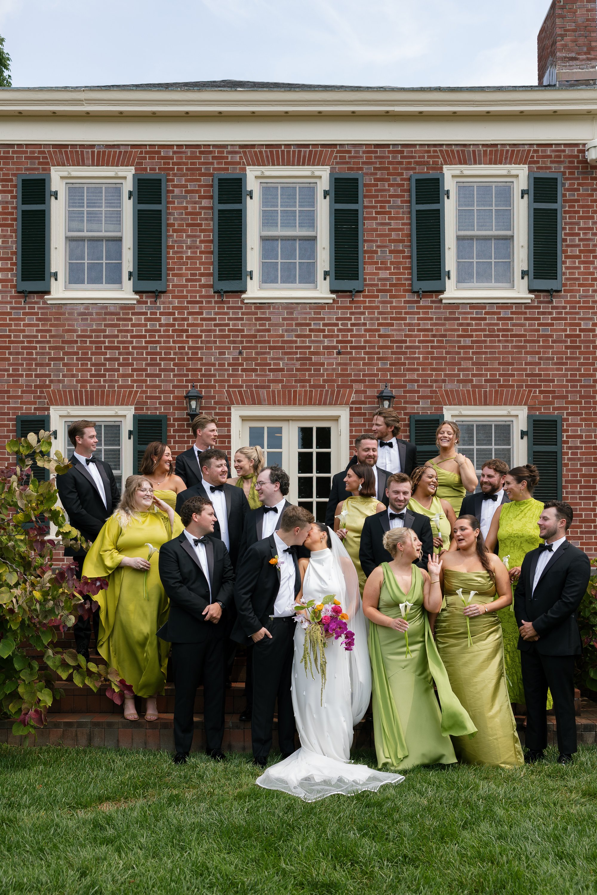 A group of wedding guests and the bride and groom standing outside a brick building with black shutters and multiple windows, during daytime with an overcast sky.