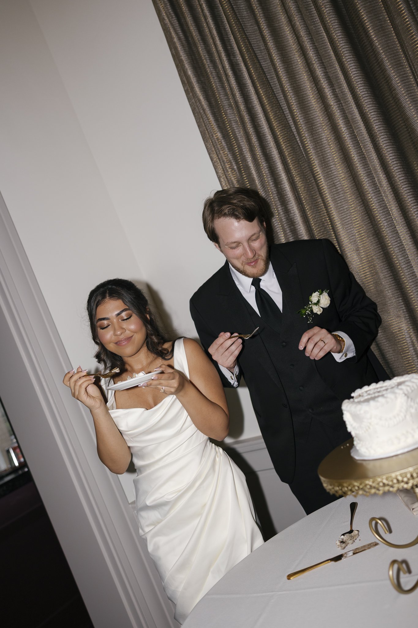 A bride and groom at a celebration, sharing a moment while cutting a wedding cake. The bride wears a white dress, and the groom in a black tuxedo with a white flower boutonniere. A cake with white frosting is on a table in front of them.