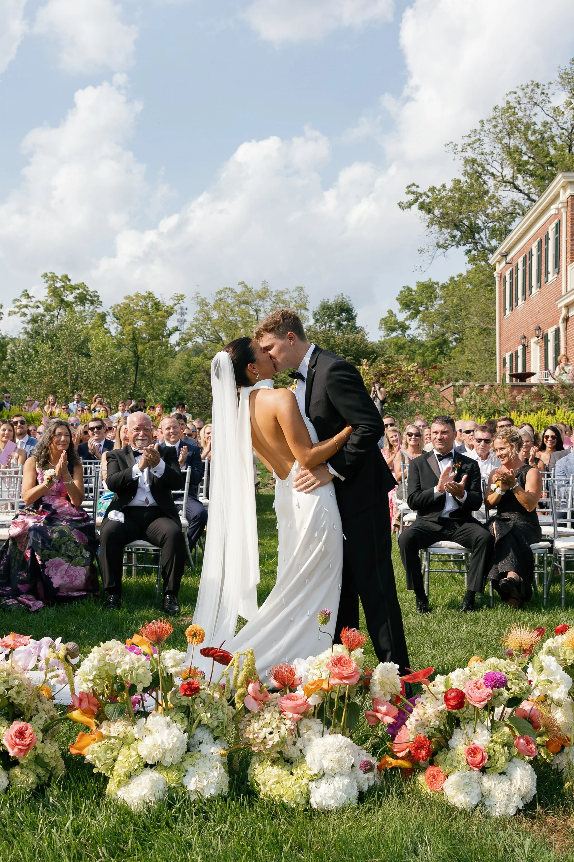 A bride and groom sharing a kiss during an outdoor wedding ceremony with guests clapping and seated in chairs, and colorful flowers arranged along the grass in the foreground.
